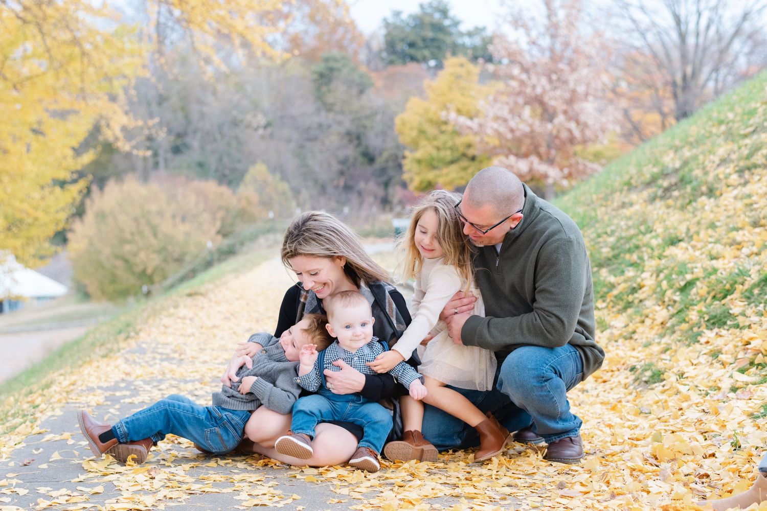 Family of four sitting on fallen autumn leaves, parents lovingly gazing at two young children outdoors.