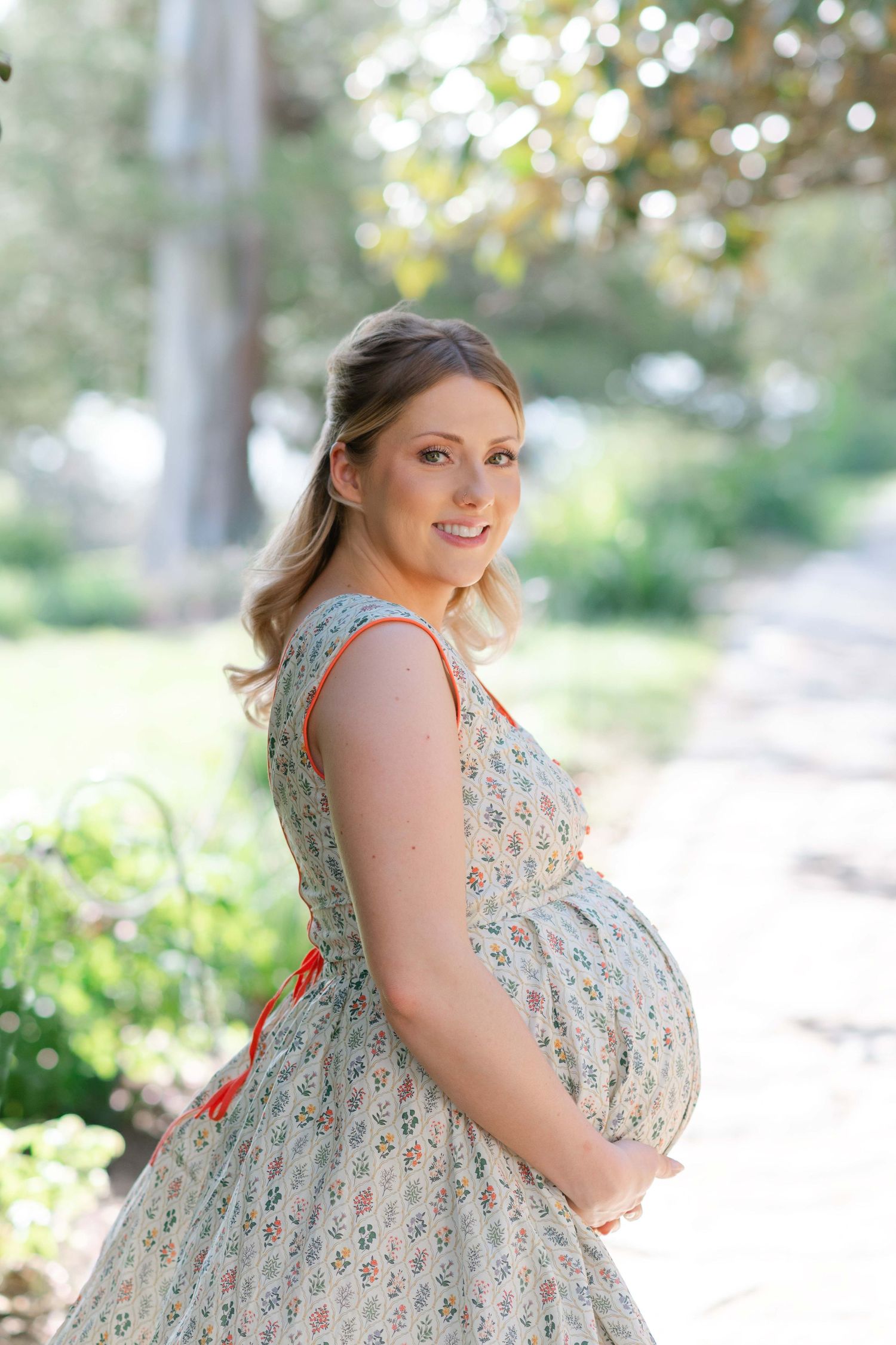 Smiling pregnant woman in floral sundress cradling baby bump outdoors in Maymont Park