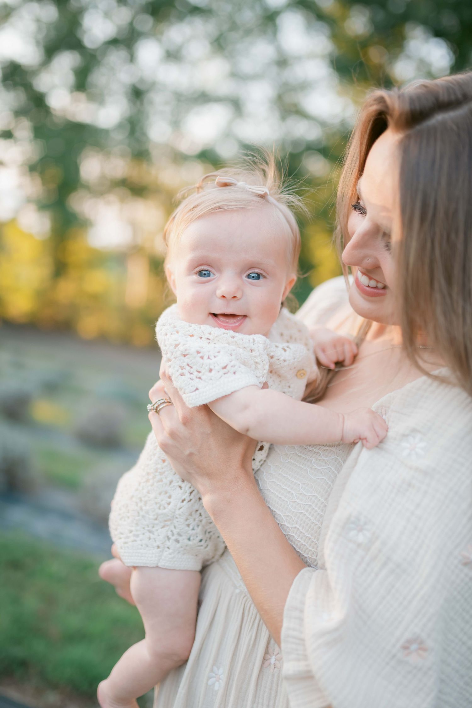 A smiling mother holds her wide-eyed baby girl in a white knit outfit outdoors in soft golden light.