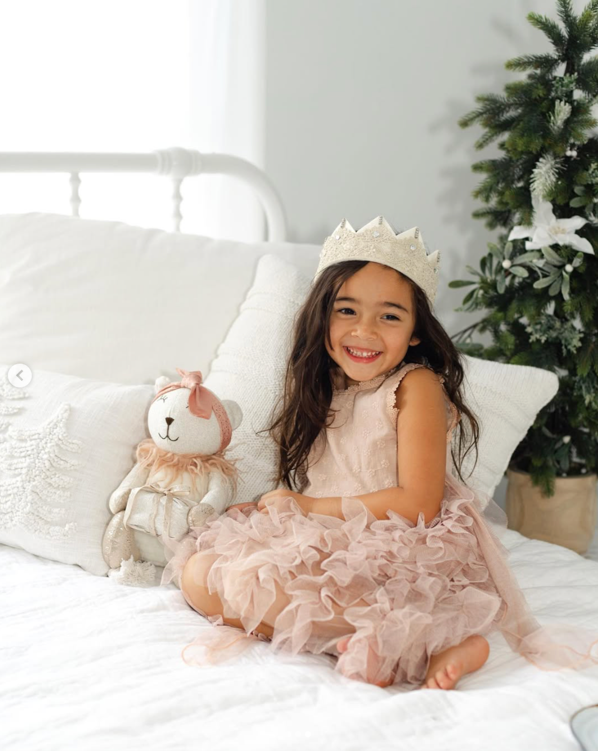A smiling girl in a pink tulle dress and crown sits on a white bed holding a stuffed bear near a Christmas tree.