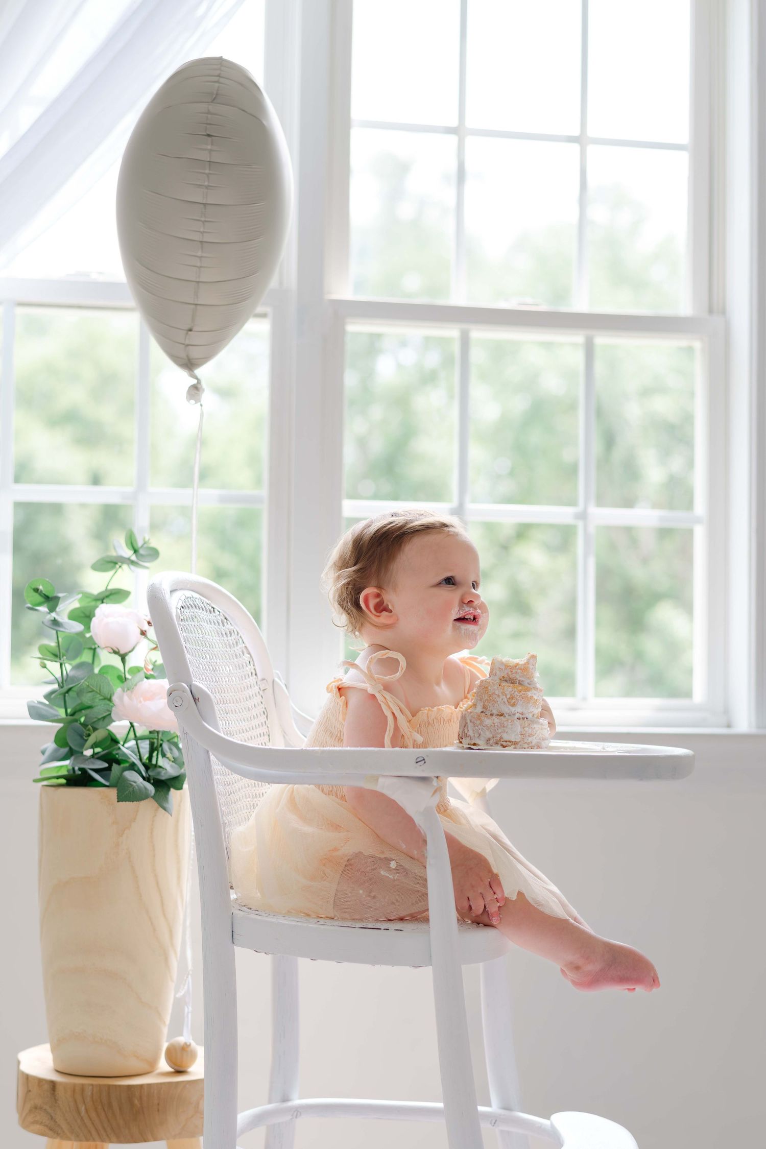 Baby girl in yellow dress sits in white high chair by window with balloon and cake for first birthday celebration.
