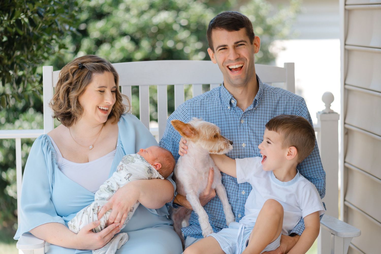 Happy family with newborn baby, young boy, and small dog sitting on porch bench, smiling outdoors.