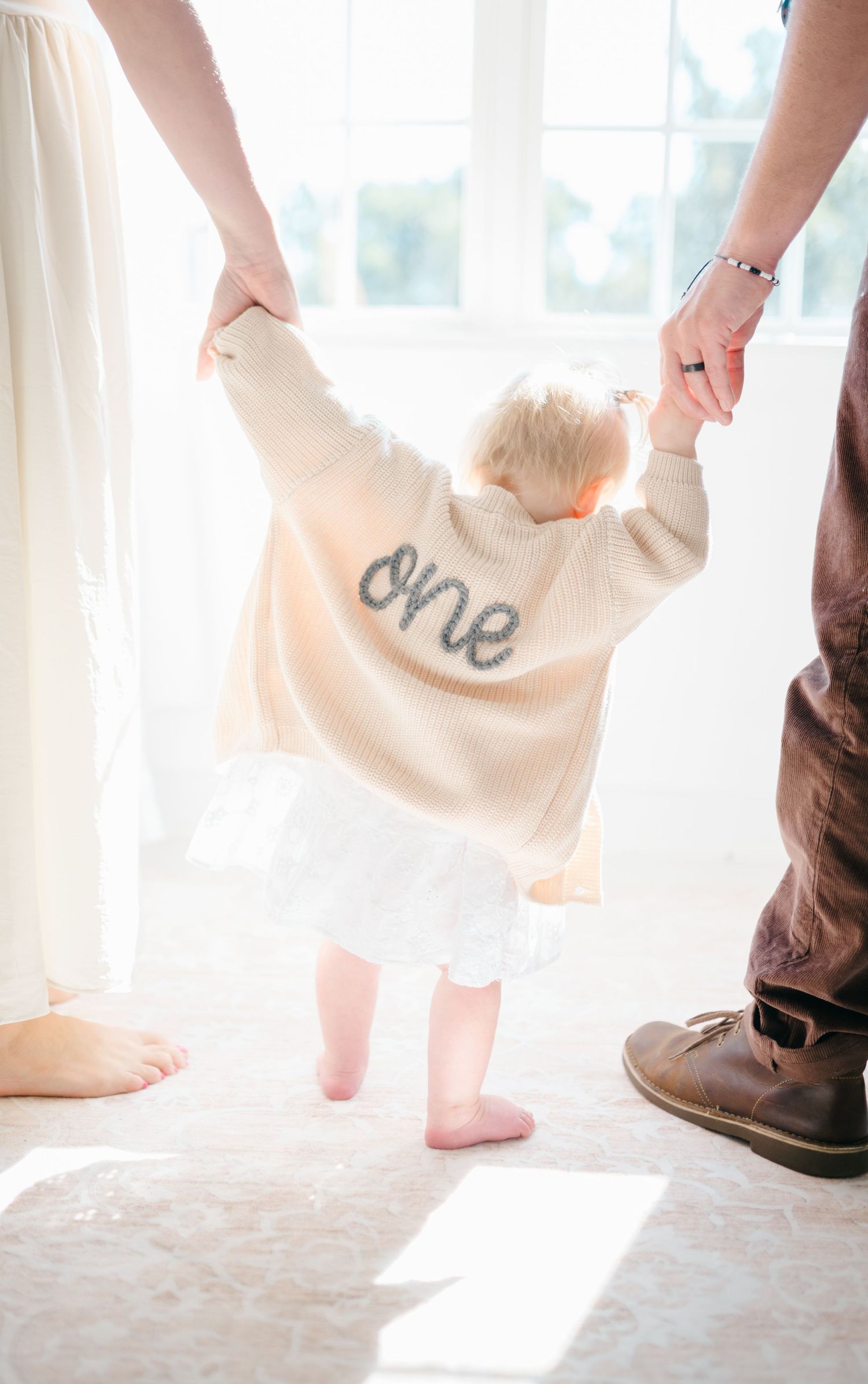 A toddler in a cream ‘one’ sweater holds parents’ hands while learning to walk in a bright, airy room.