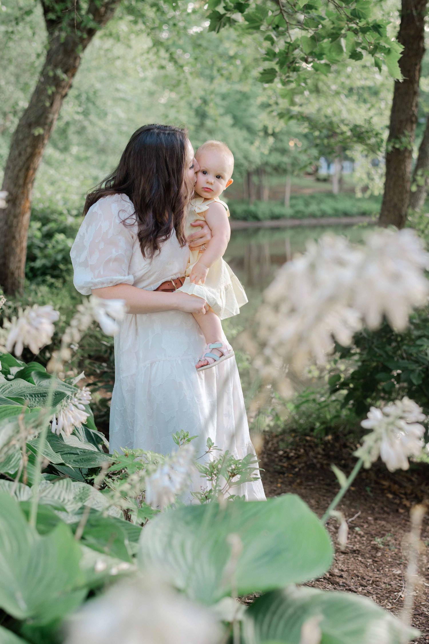 A mother in a white dress kisses her baby in a lush green garden with white flowers and trees.