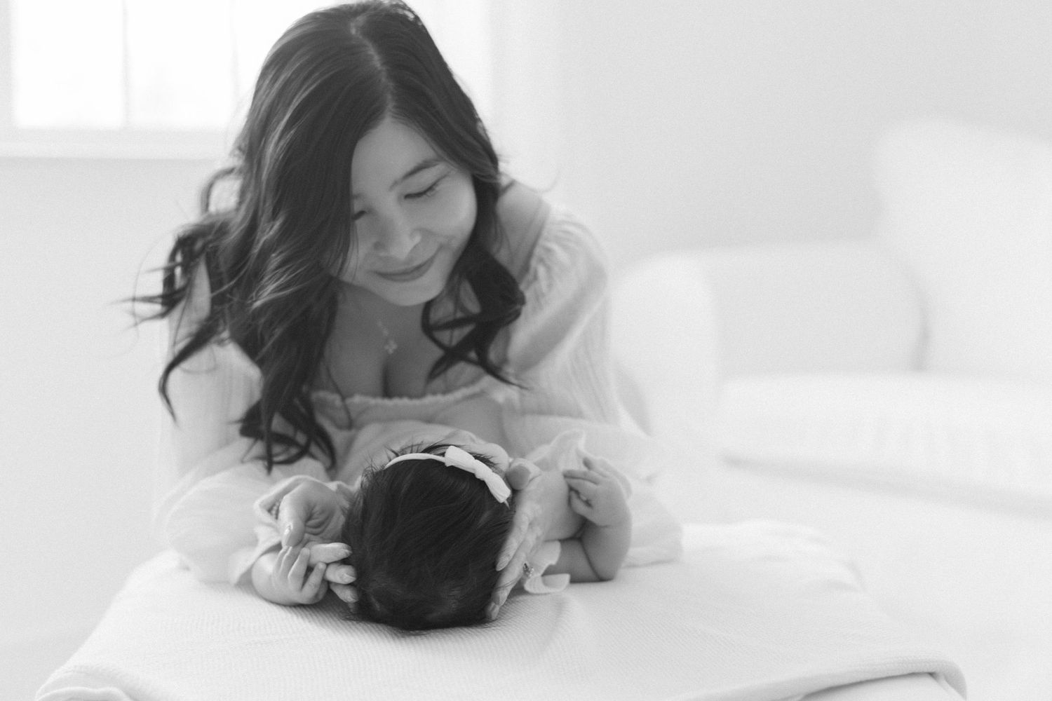 Black and white photo of a smiling Asian mother tenderly gazing at her newborn baby on a white bed.
