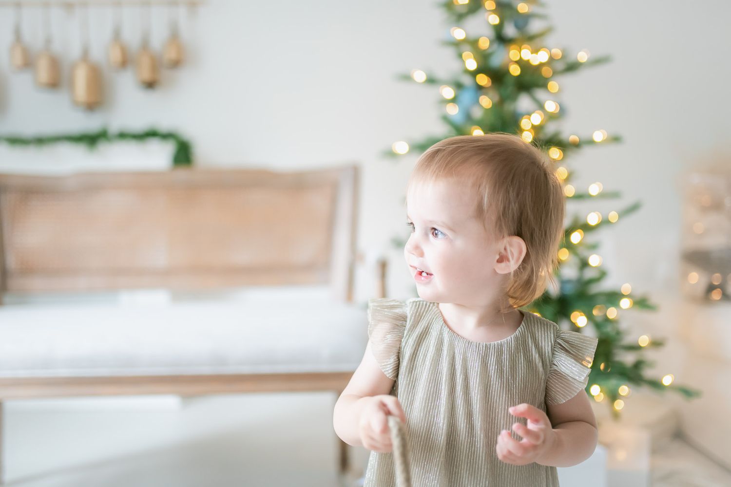 A small child in a gray dress plays near a decorated Christmas tree in a bright, modern living room.