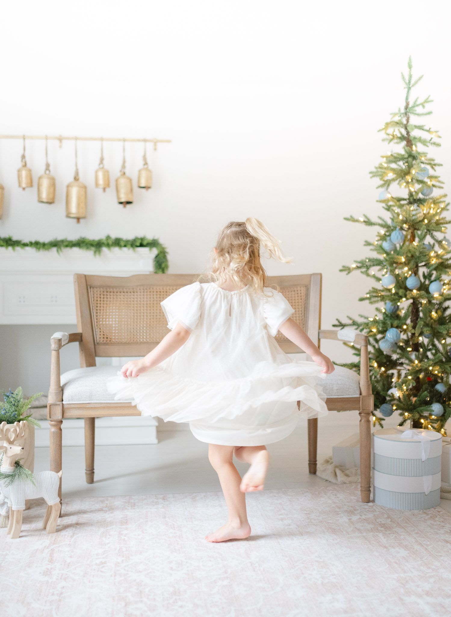 A child in a white dress twirls and dances joyfully in front of a decorated Christmas tree in a bright white room.
