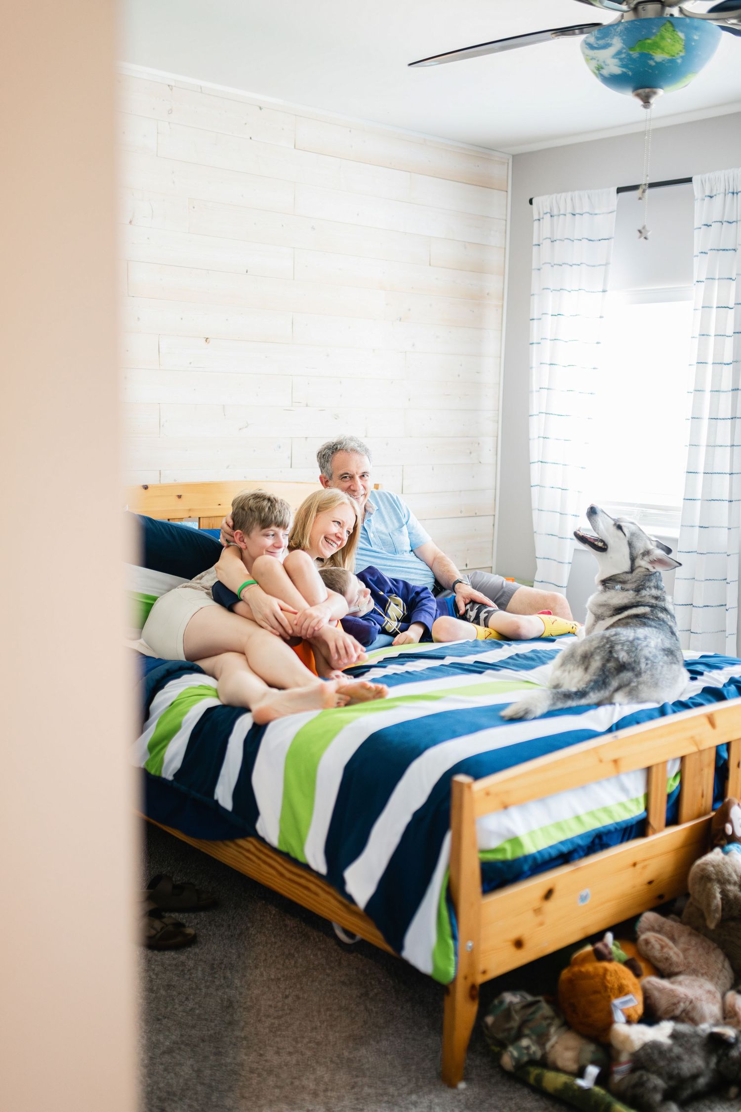 A man and two children sit on a striped bed with a Husky dog in a cozy bedroom with shiplap walls.
