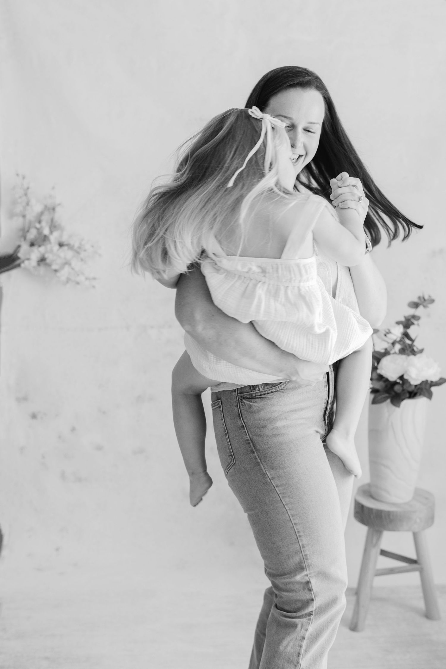 Black and white photo of a woman giving a laughing girl a piggyback ride indoors with flowers in background.