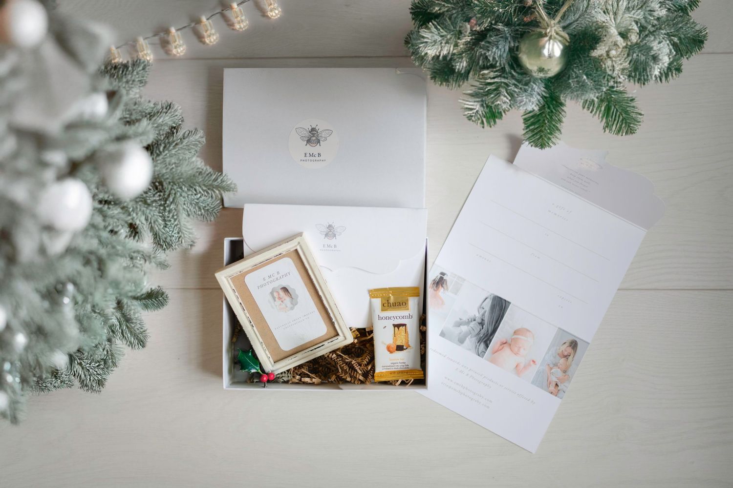 White gift box with honey jar and photo cards arranged with silver Christmas ornaments and evergreen branches on light surface.
