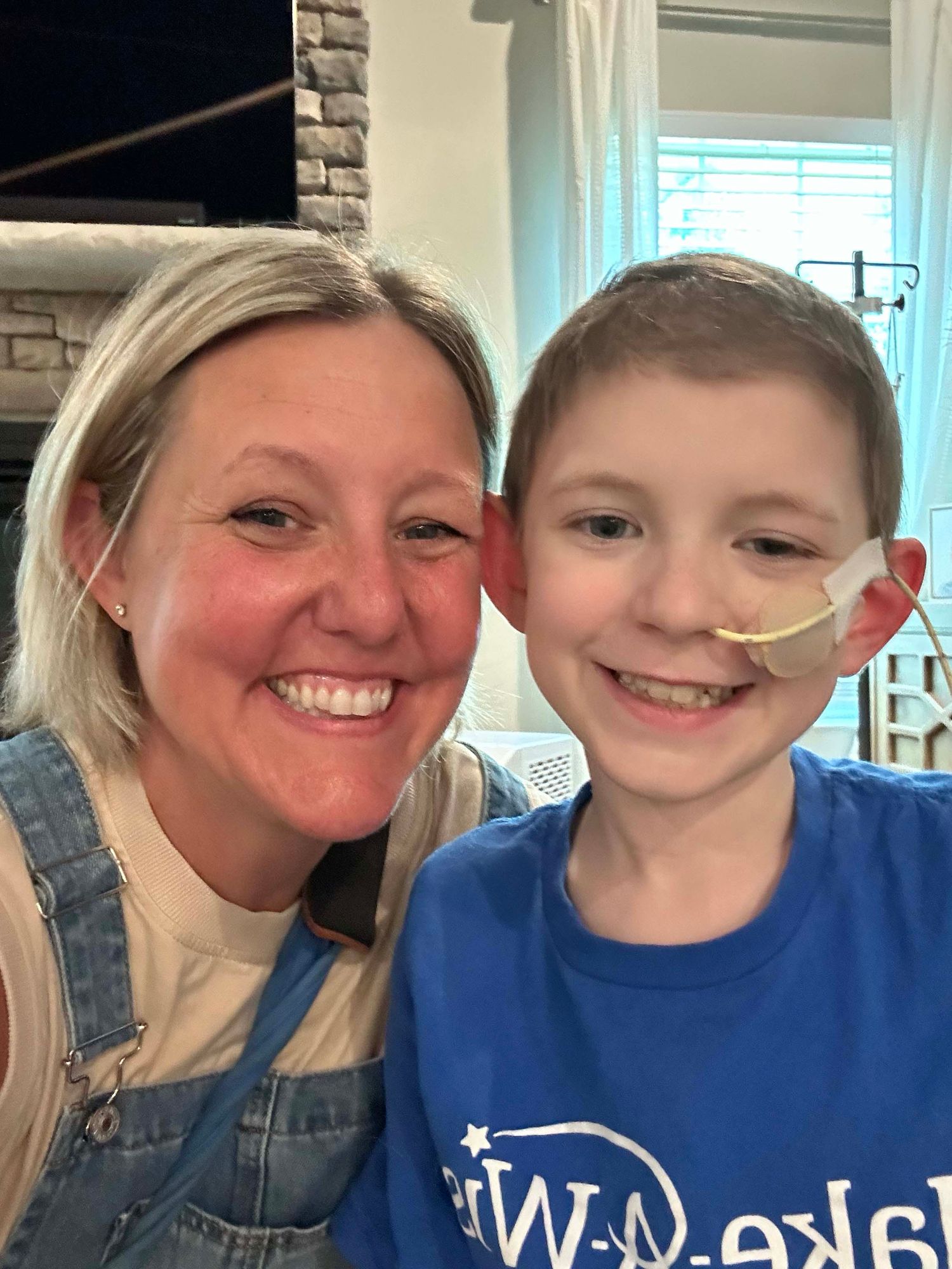A smiling woman and young boy with a feeding tube pose together for a selfie indoors.