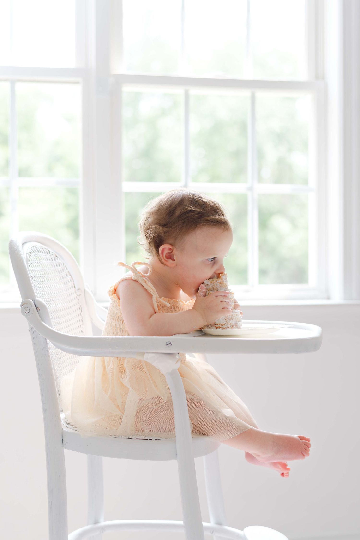 Baby girl in peach dress eating cake in white high chair by bright window.