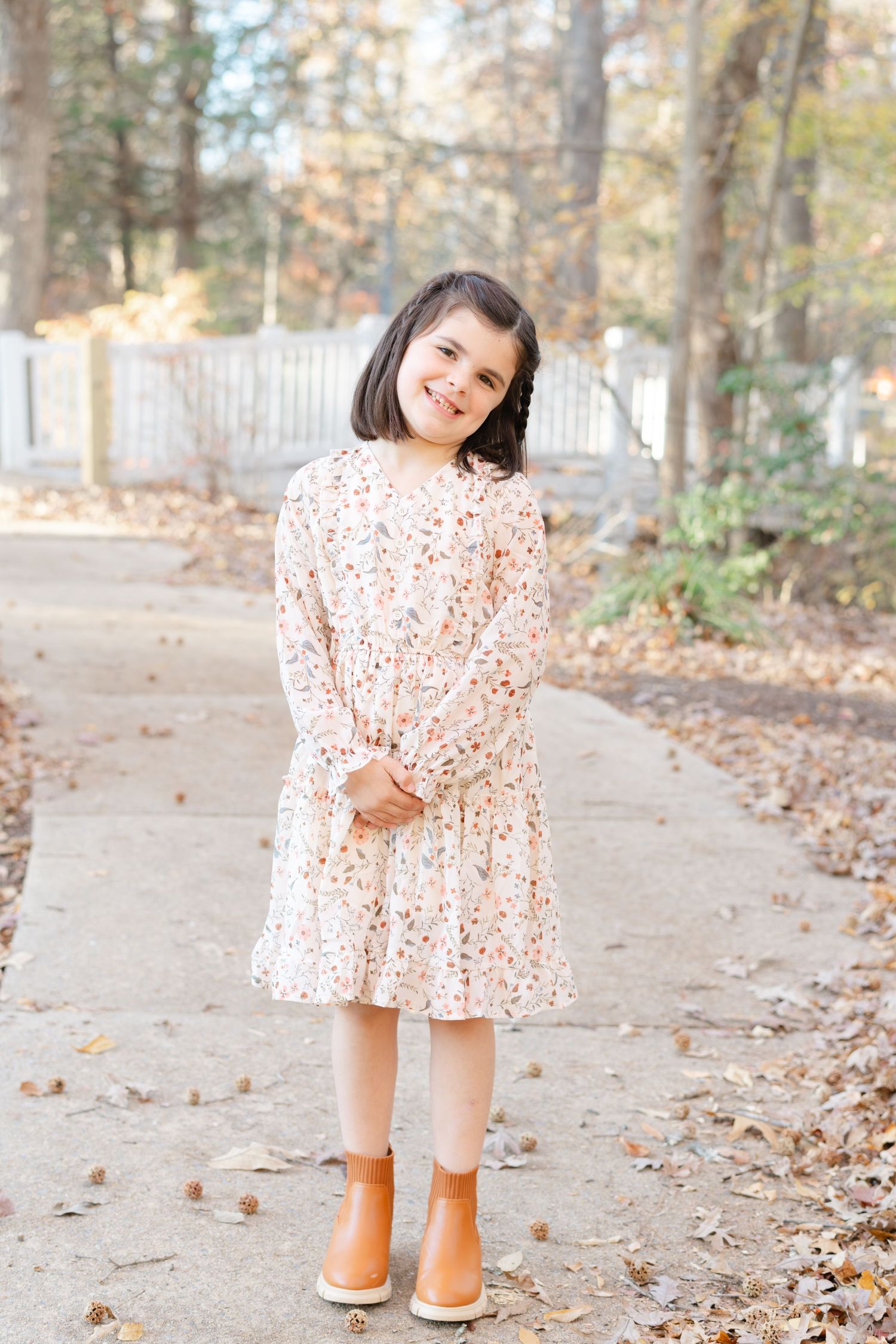Young girl in floral dress and orange boots smiling on a garden path surrounded by autumn leaves.