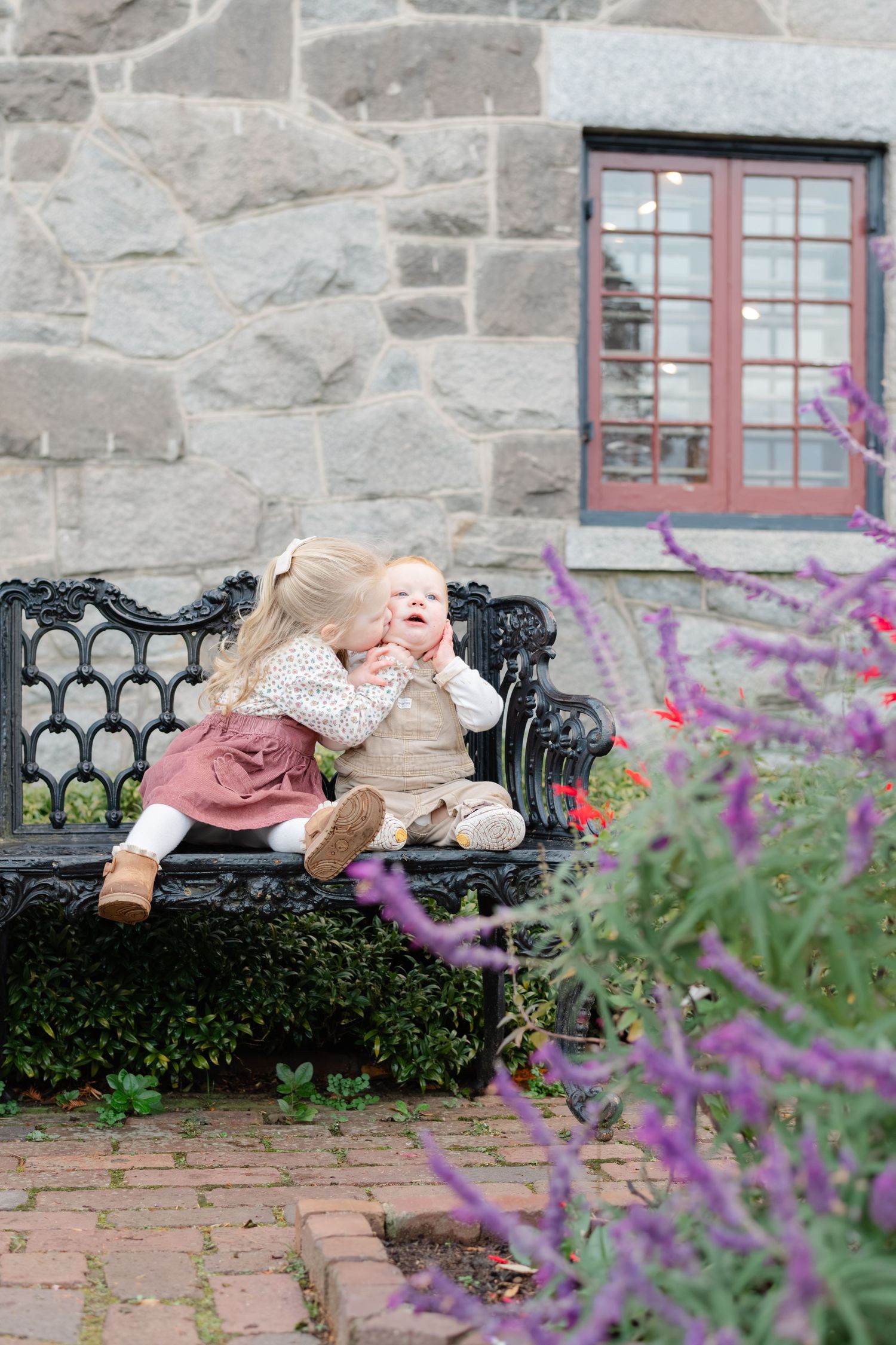 Two young children sit on a vintage black bench, with the older girl hugging her baby sibling outdoors.