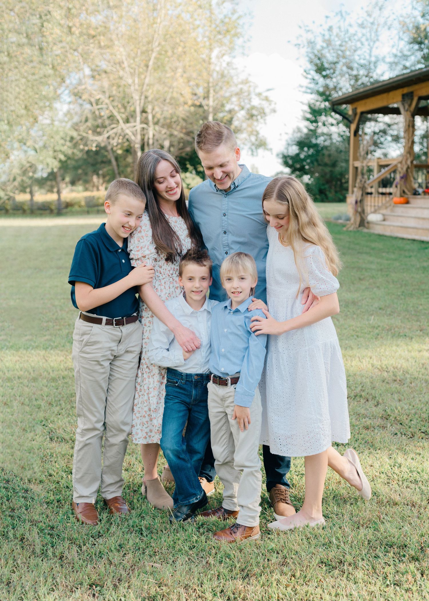 A happy family of six poses outdoors on green grass, smiling together near trees and a wooden gazebo.