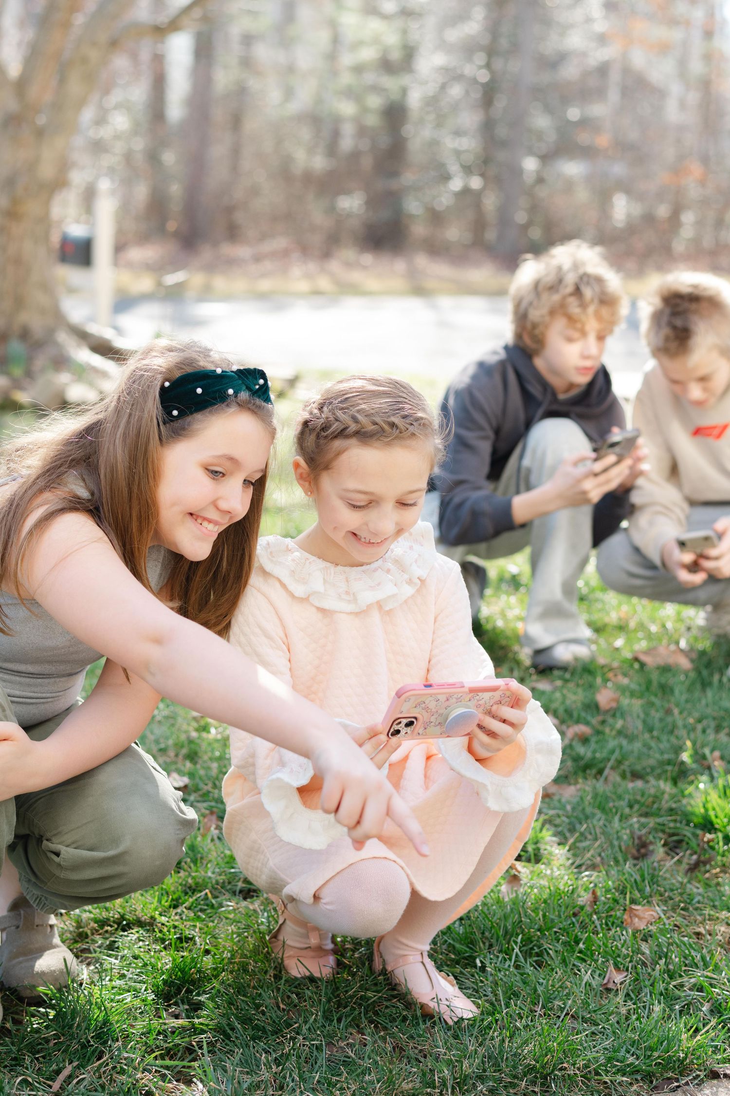 People sitting on grass in a wooded area looking at mobile phones and devices on a sunny day.