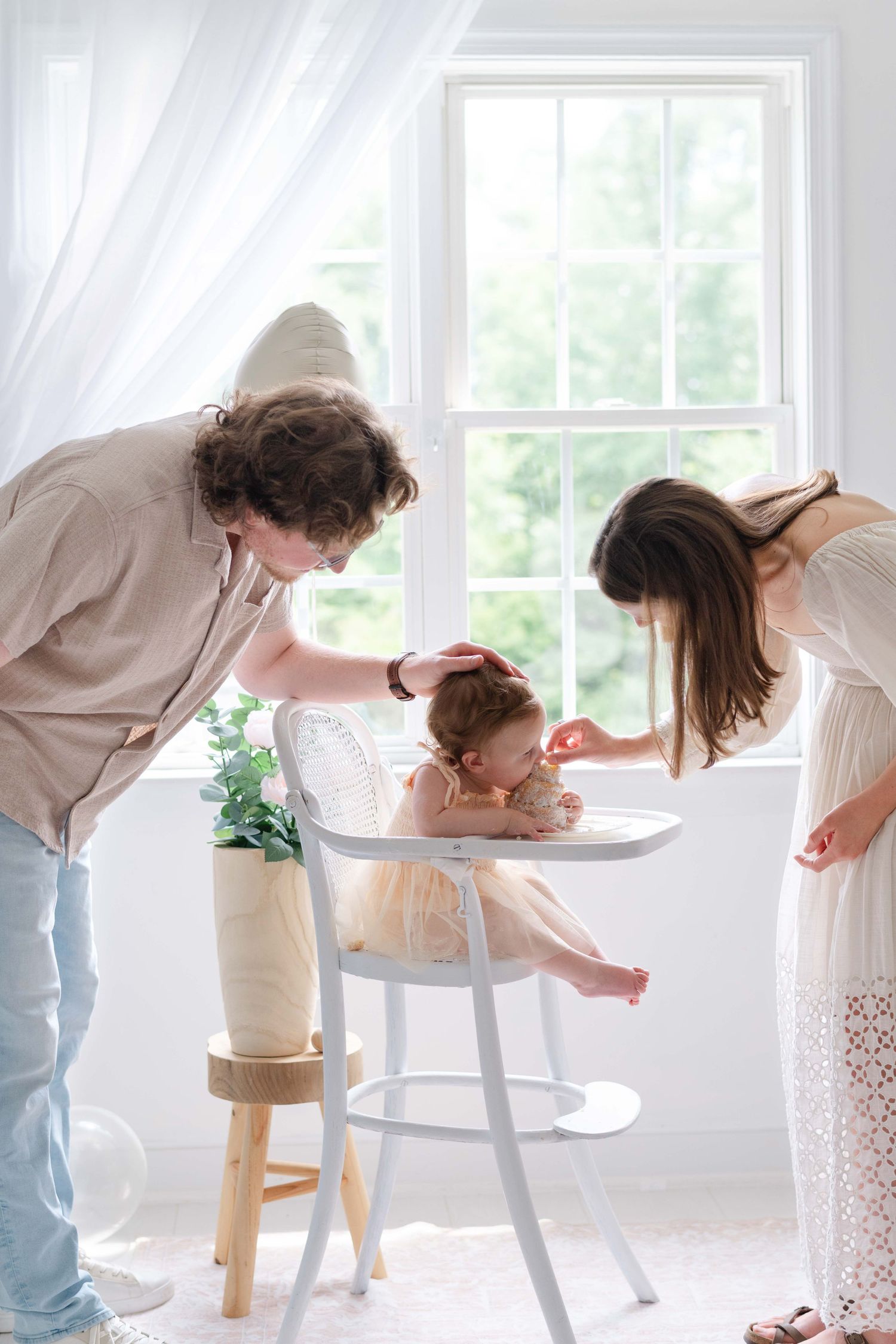 Two parents lovingly tend to their baby sitting in a white high chair by a bright window in a light-filled room.
