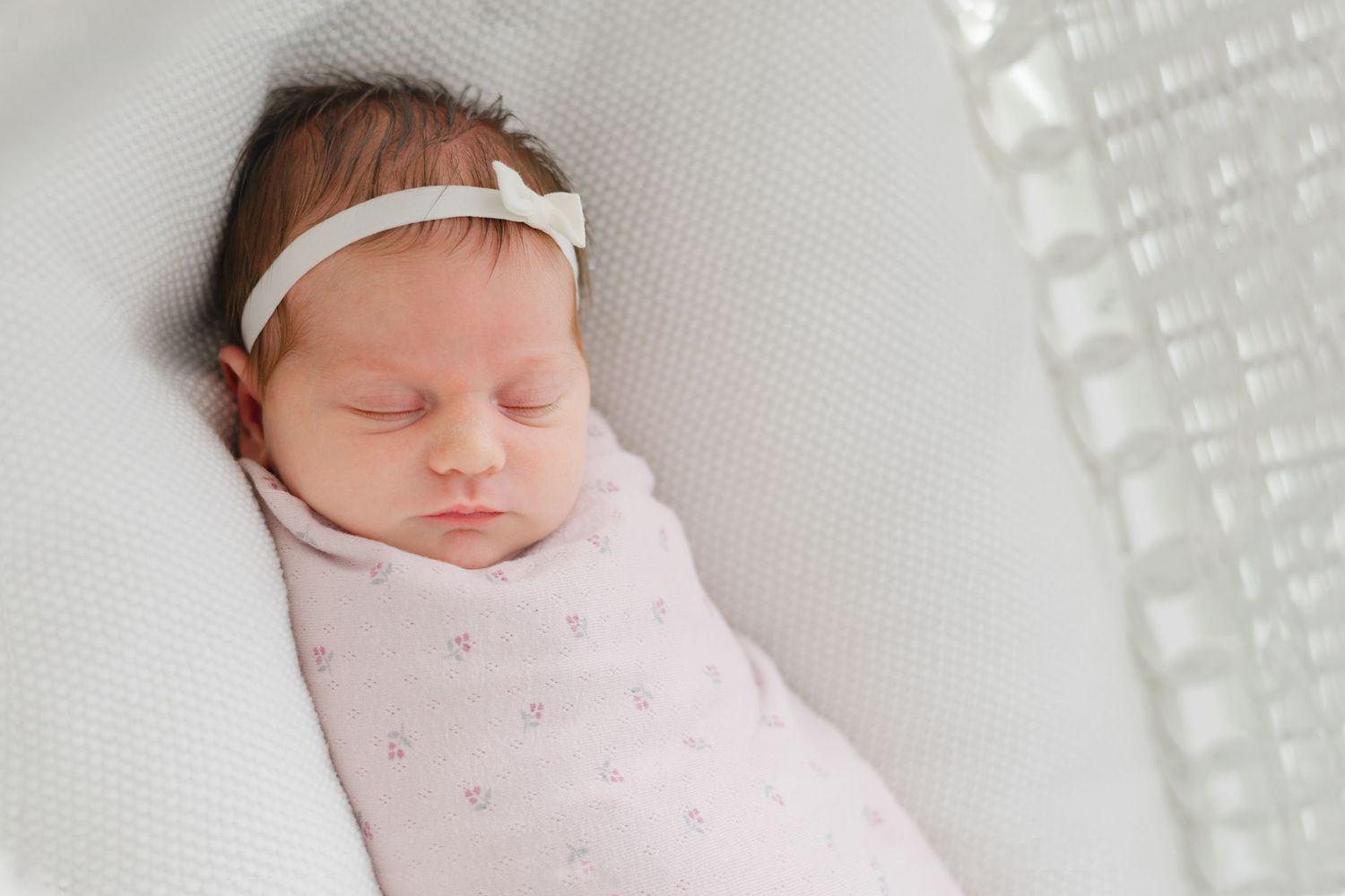 Newborn baby girl swaddled in pink blanket sleeping peacefully in white bassinet wearing white bow headband.