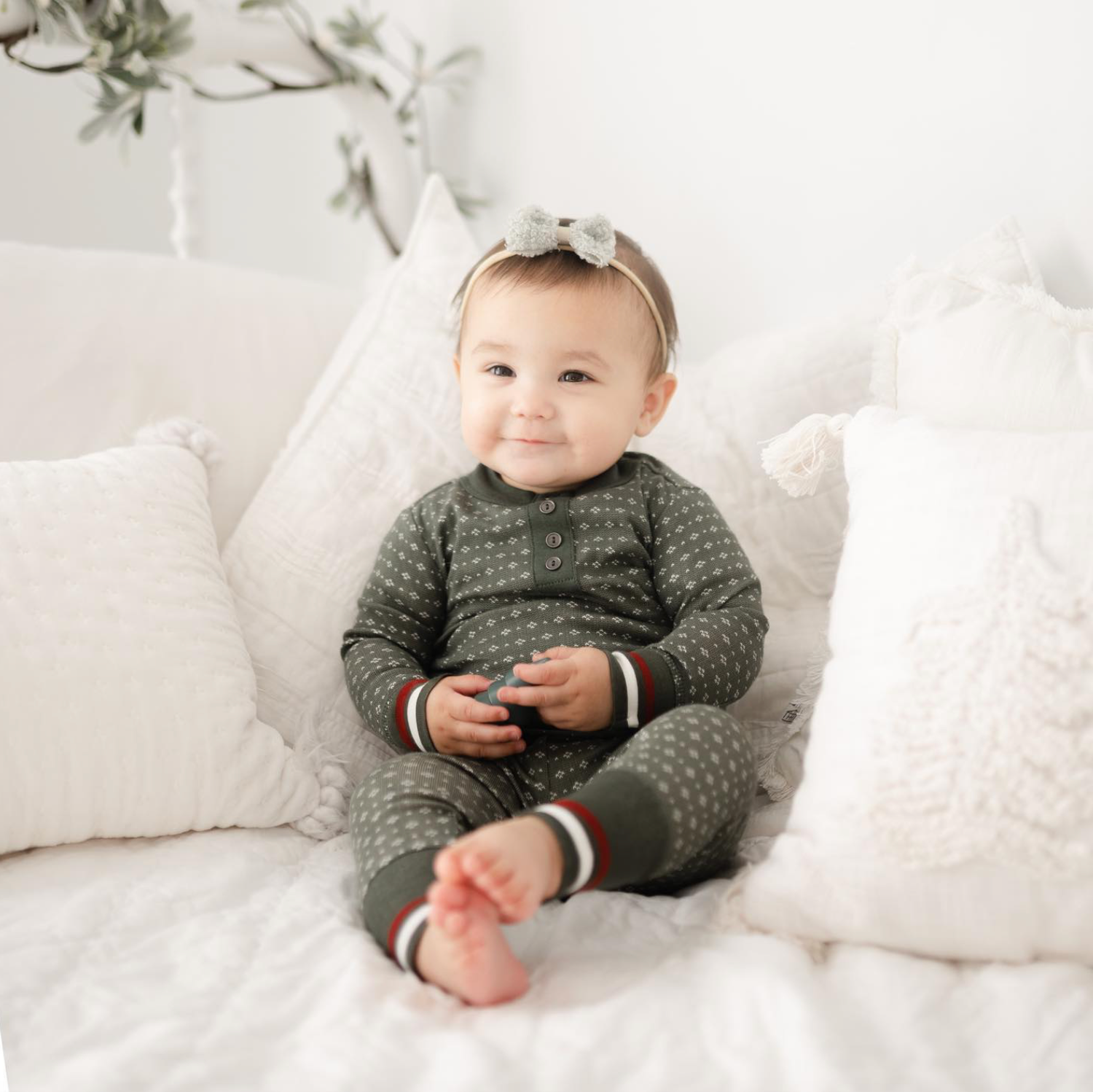 Smiling baby in green polka dot pajamas with a bow headband sits on white cushions.