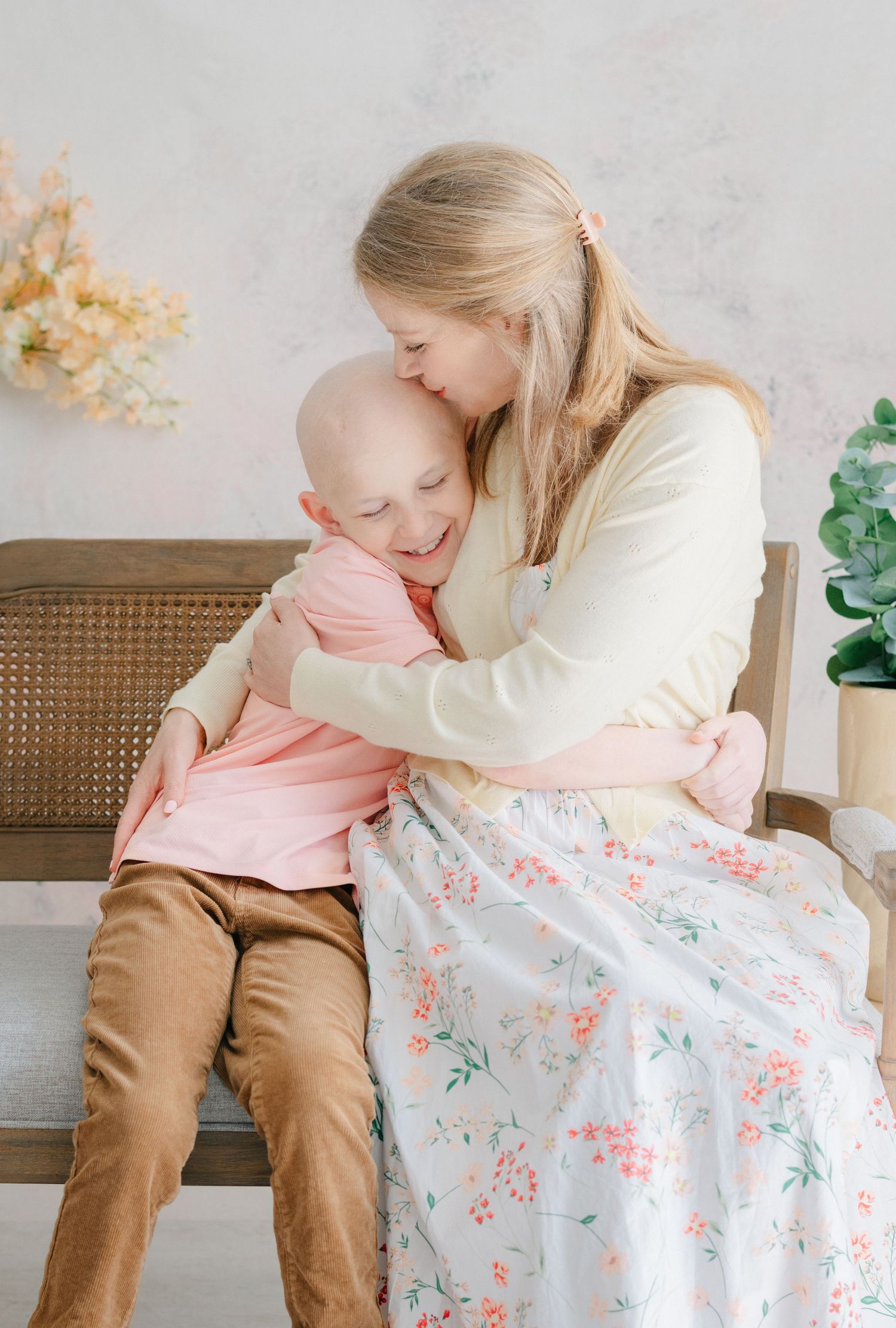 A mother tenderly hugs her young child with hair loss, both smiling warmly while seated on a wooden bench.