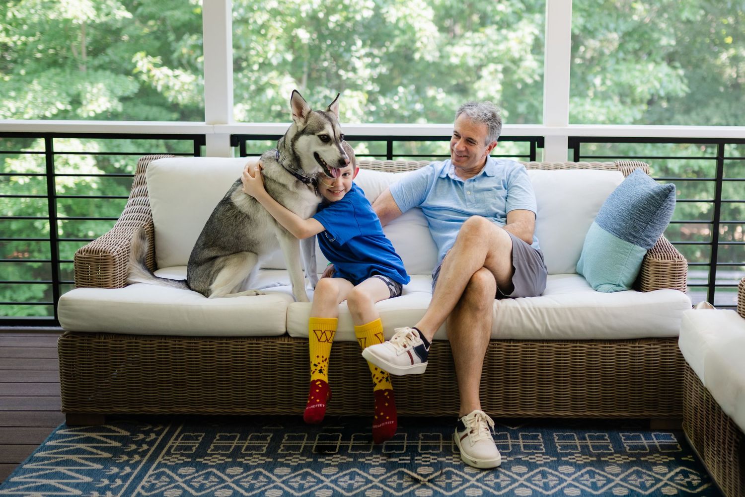 A boy and his father sit on a patio couch with a Husky dog in a screened porch surrounded by trees.