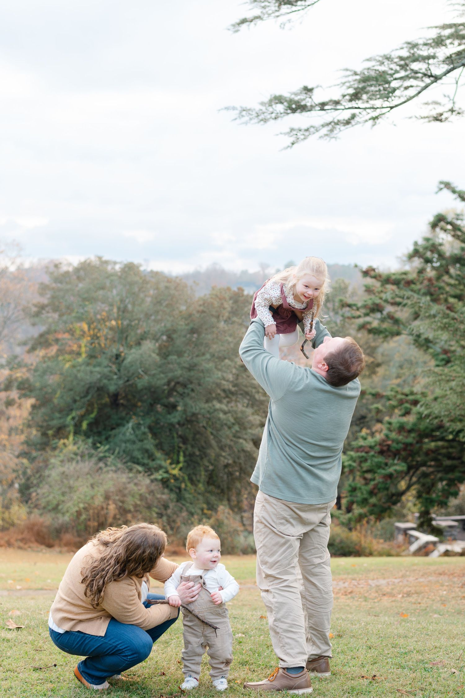 Family of four enjoying autumn outdoors, parents playing with two young children on green grass with fall trees behind.