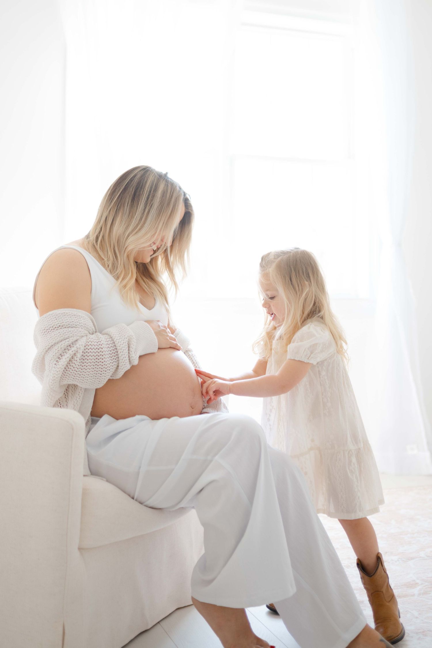 Pregnant woman sitting in white chair with young blonde girl touching her bare baby bump in bright room.