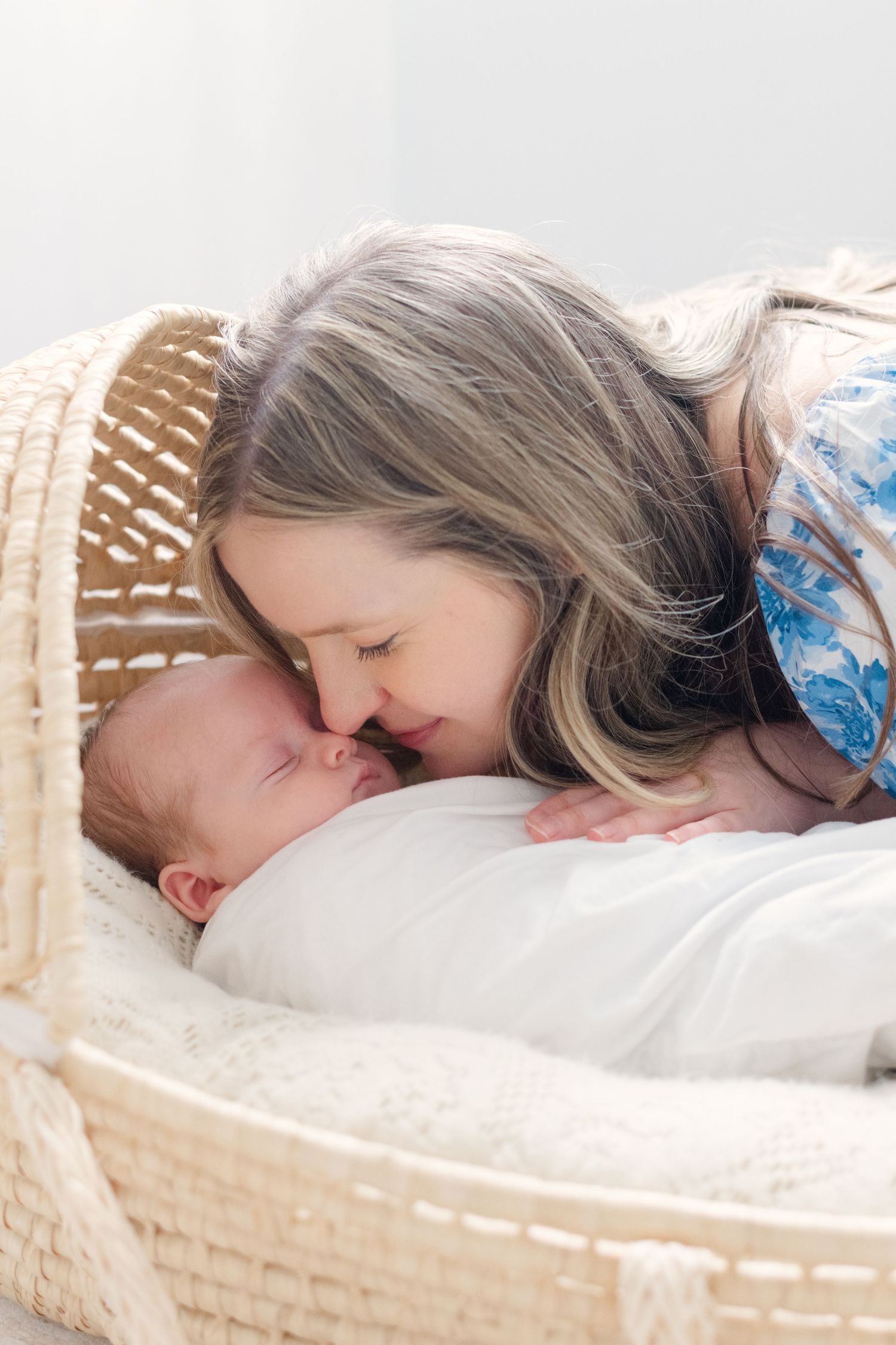 A mother tenderly touches noses with her swaddled newborn baby lying in a wicker Moses basket.