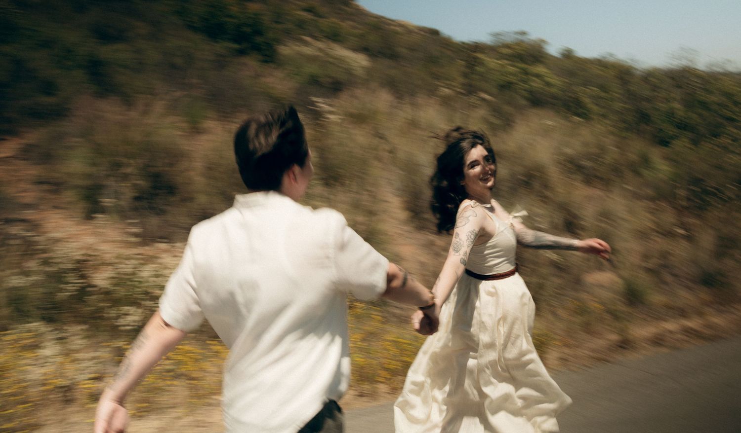 A loving couple runs and spins joyfully together on a scenic hillside path while wearing white outfits.