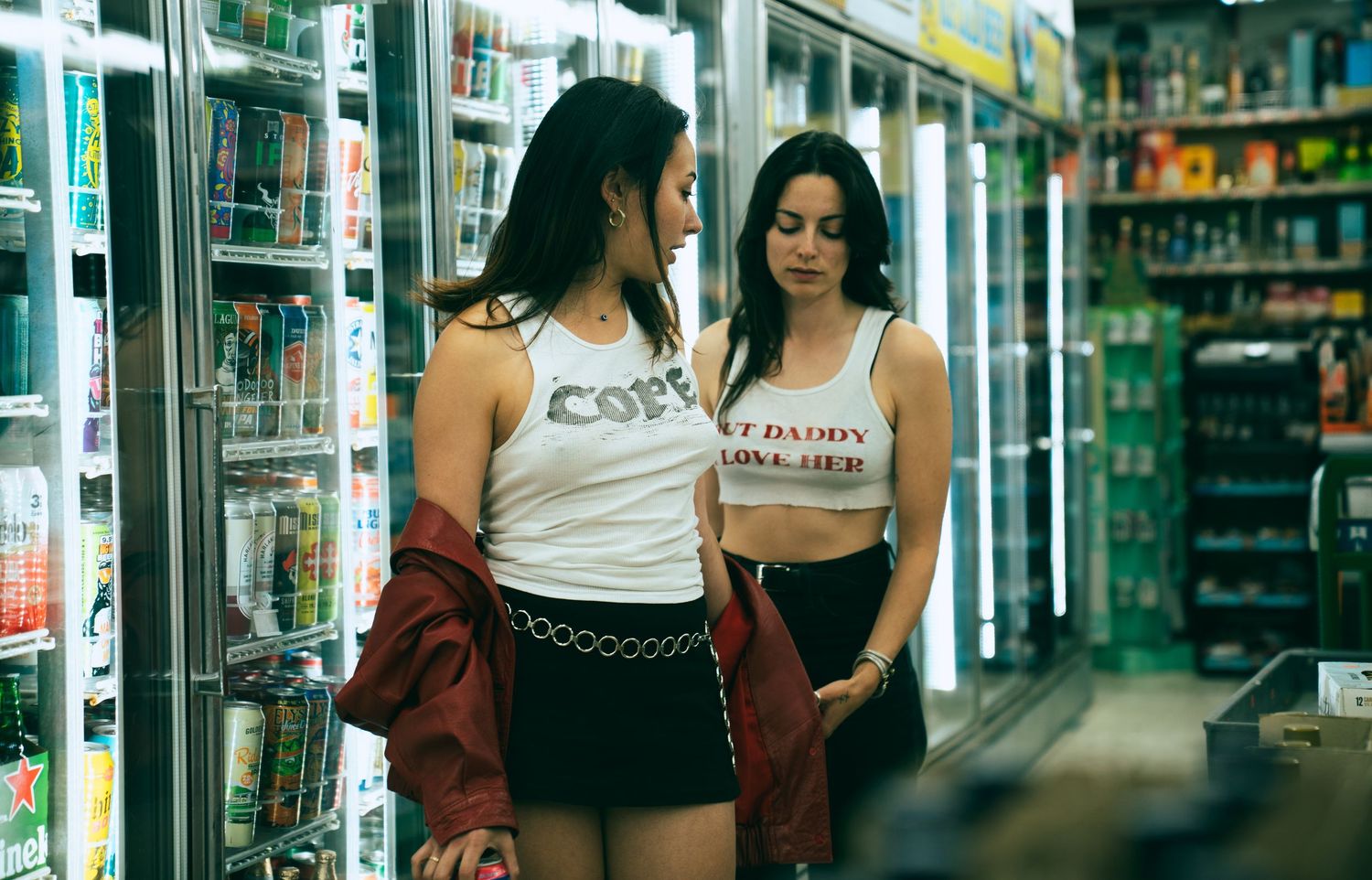 Two customers browse products in convenience store cooler aisle with glass refrigerator doors and merchandise displays.