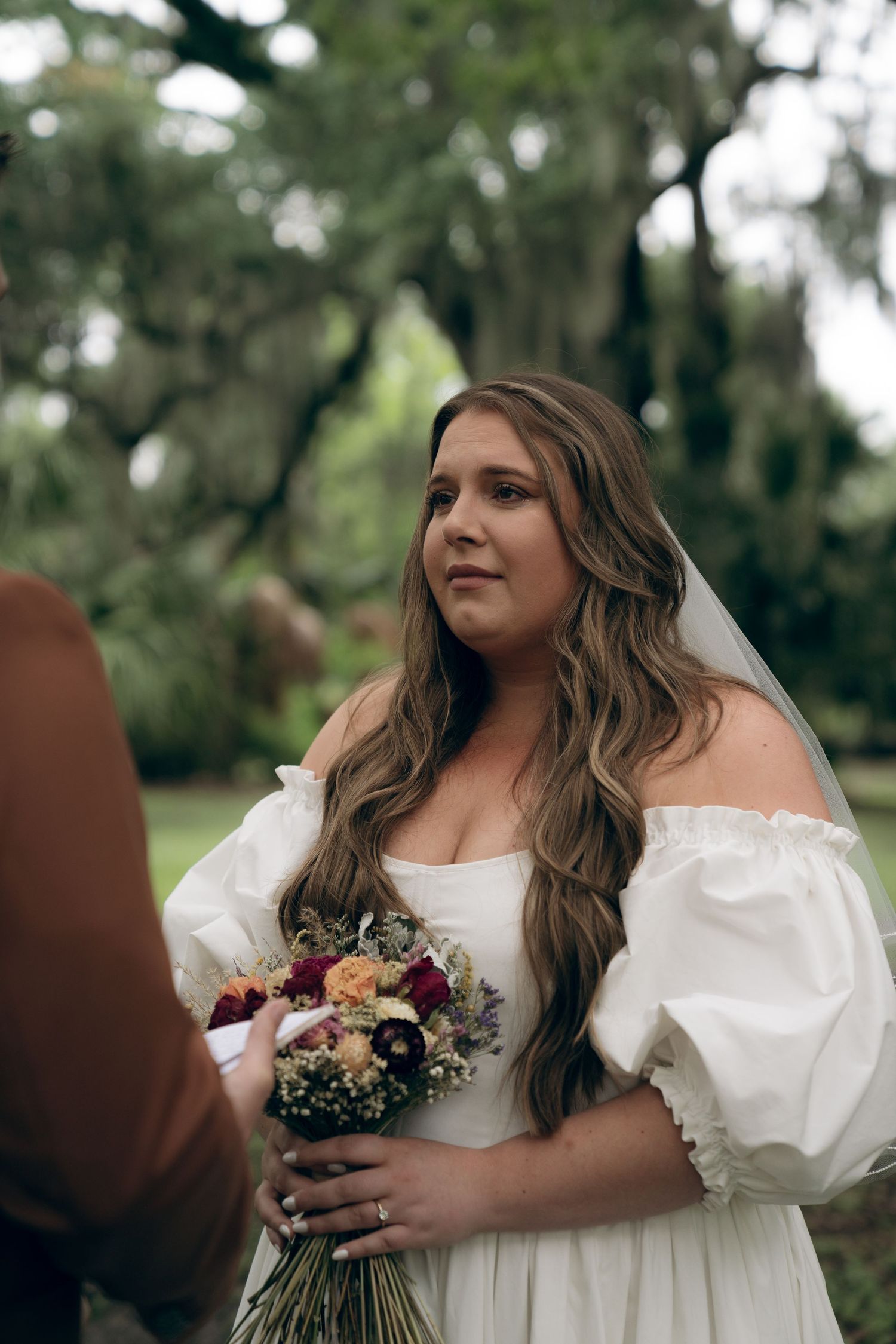A bride in an off-shoulder white wedding gown holds a colorful bouquet outdoors with trees in the background.
