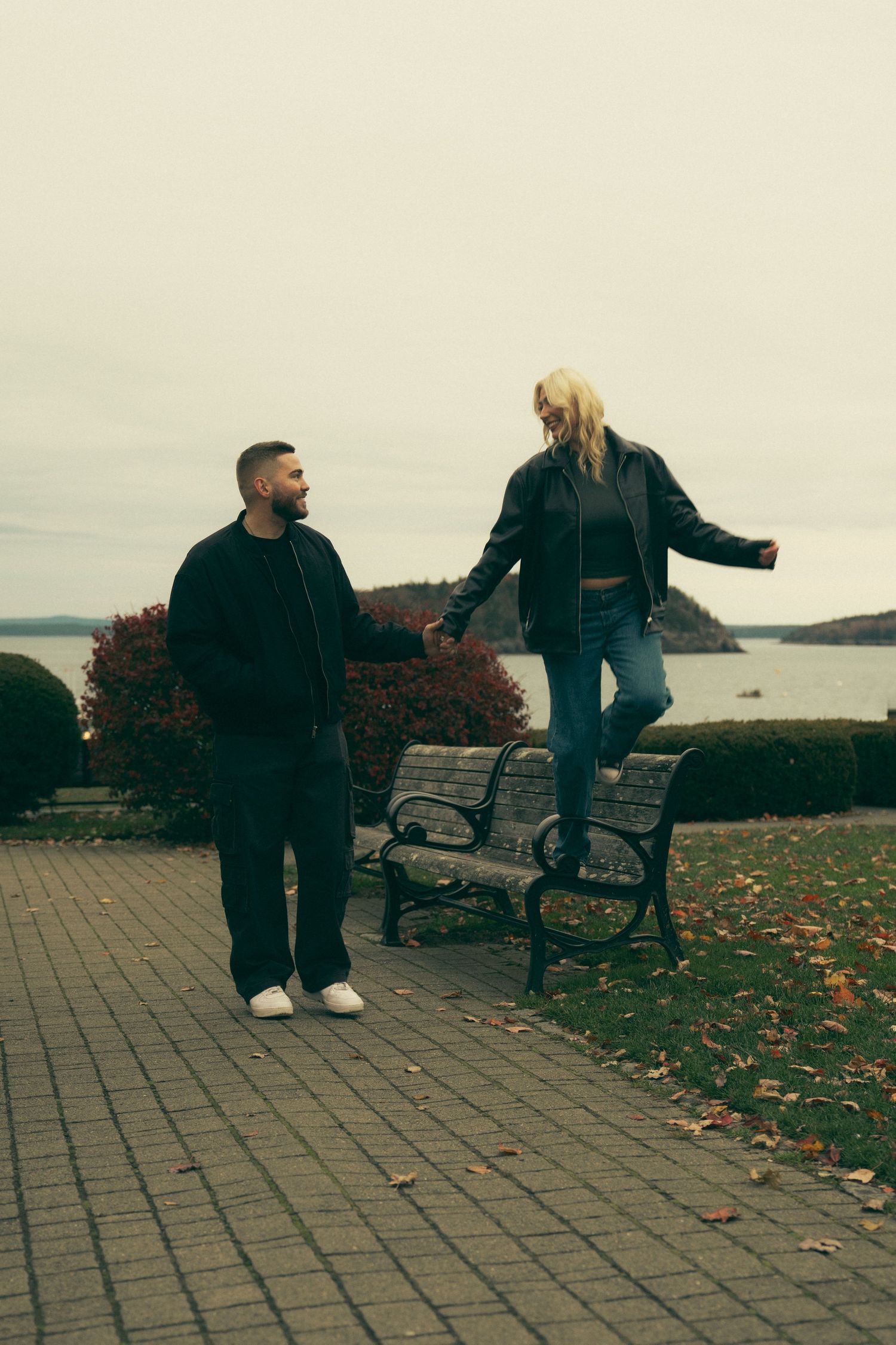 Two people share a playful moment on a park bench overlooking a coastal landscape on an autumn day. Couple in Bar Harbor Maine