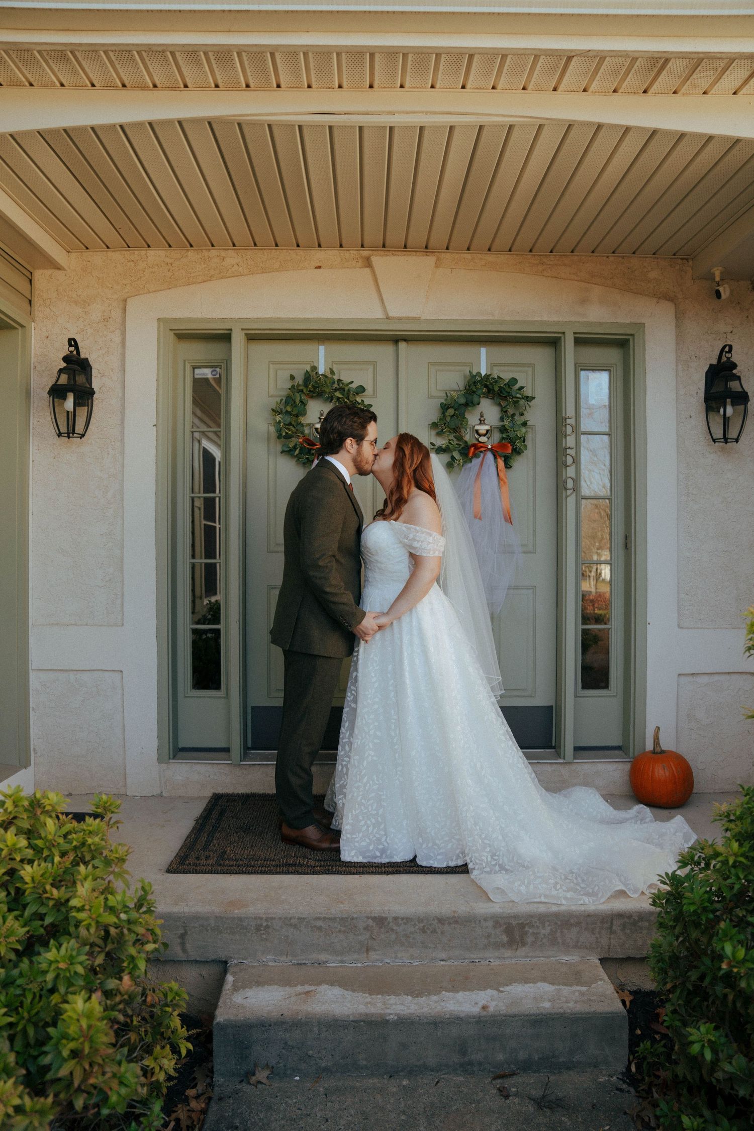 Wedding couple embracing on front porch decorated with winter wreaths and pumpkins.