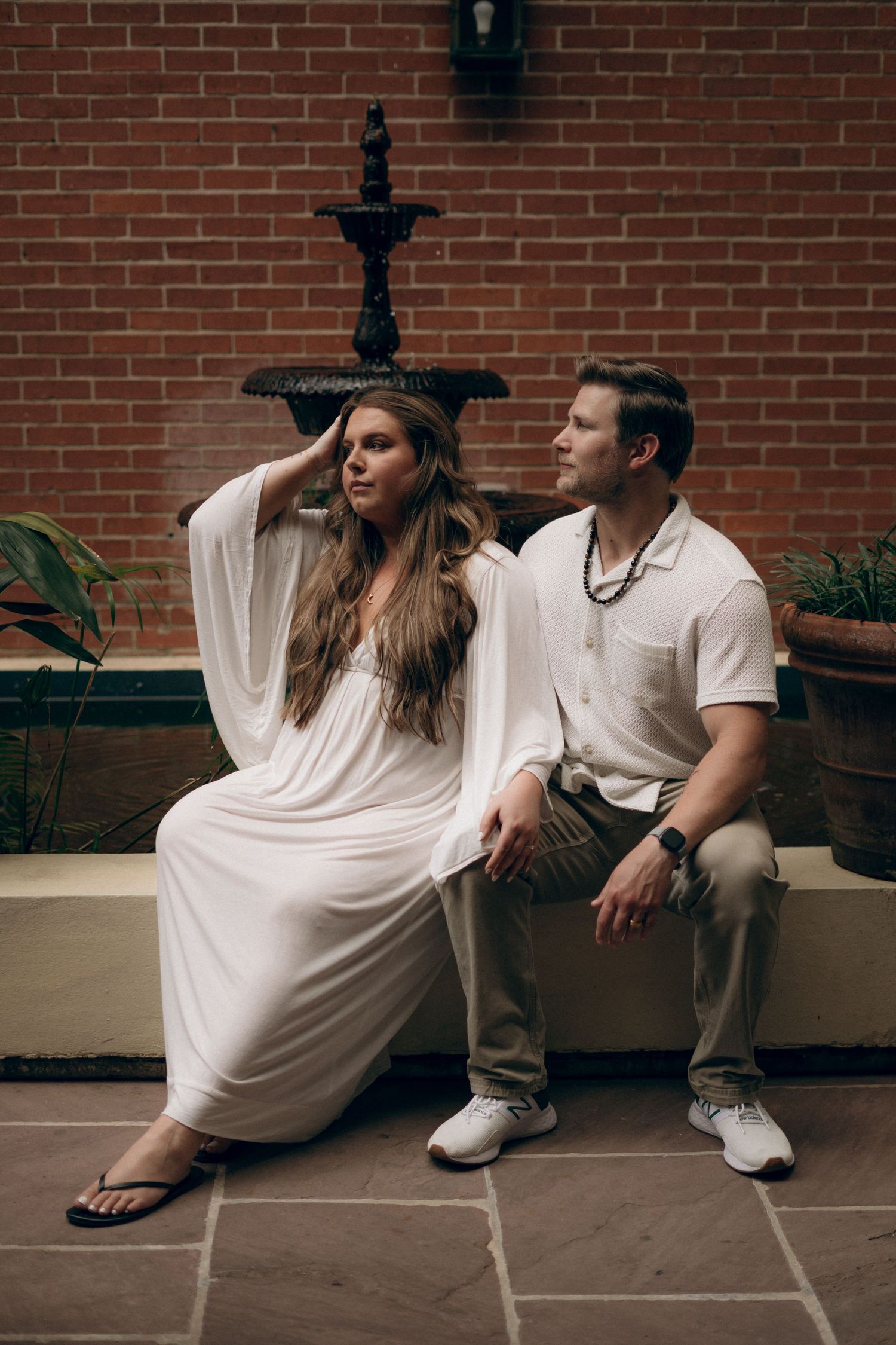 A couple sits together on a stone bench against a brick wall with a decorative fountain in a serene courtyard setting.