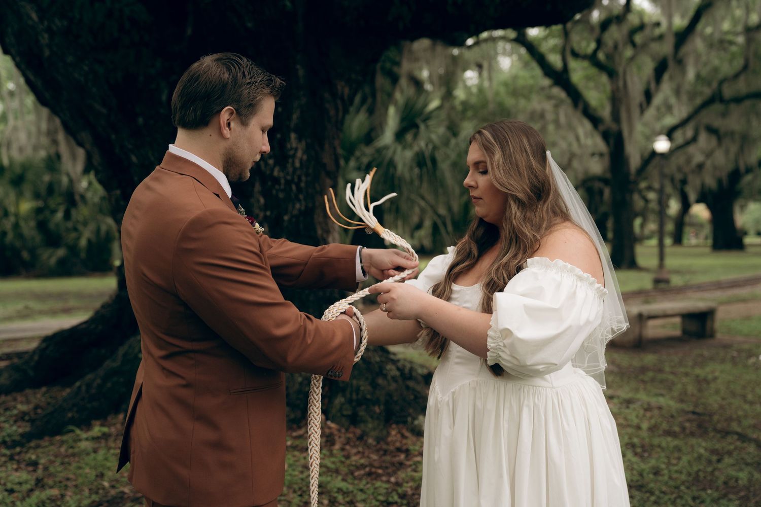 A couple in formal attire shares an intimate moment during an outdoor wedding ceremony under trees.