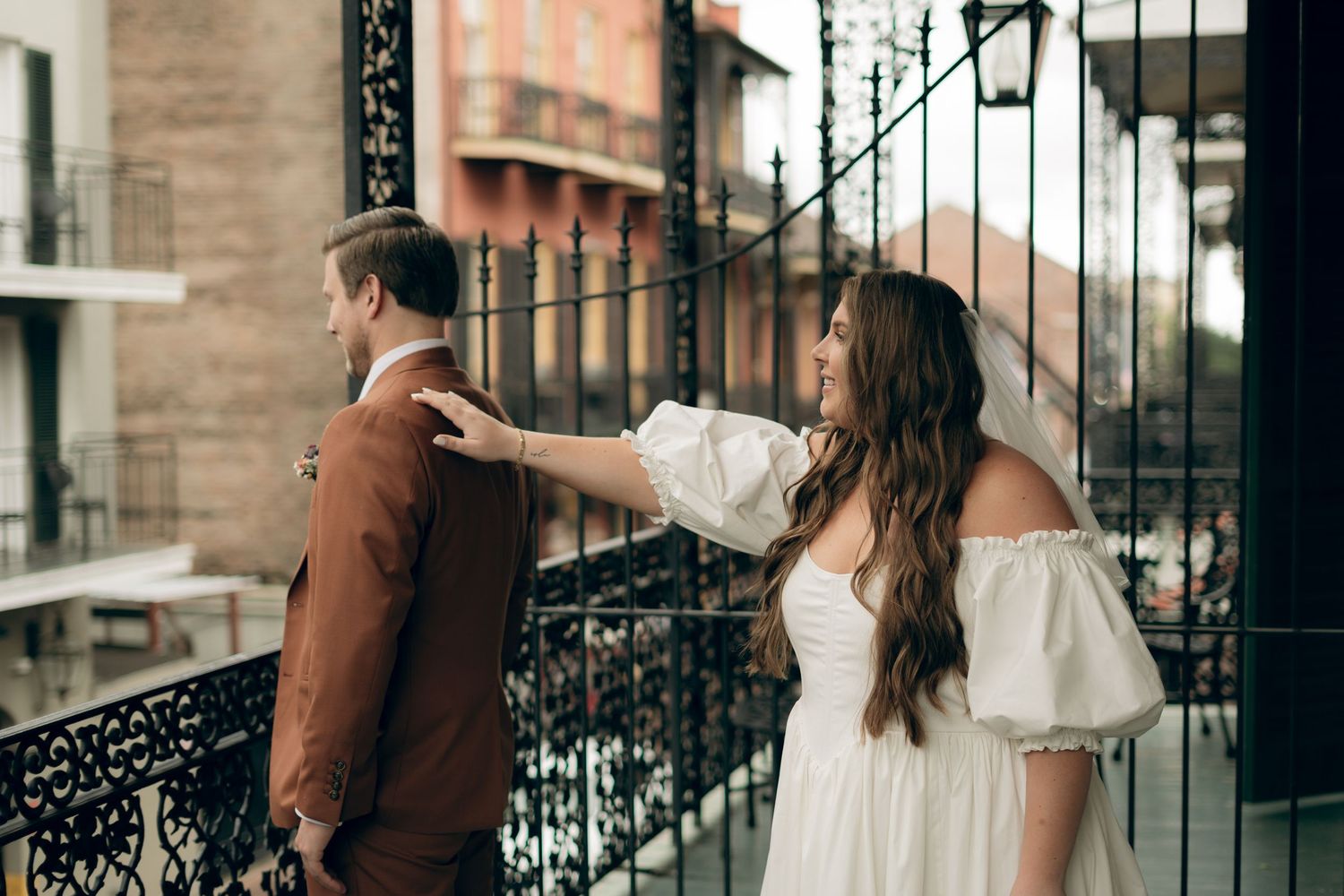 Couple poses on ornate iron balcony, person in white flowing dress reaching toward person in brown jacket.
