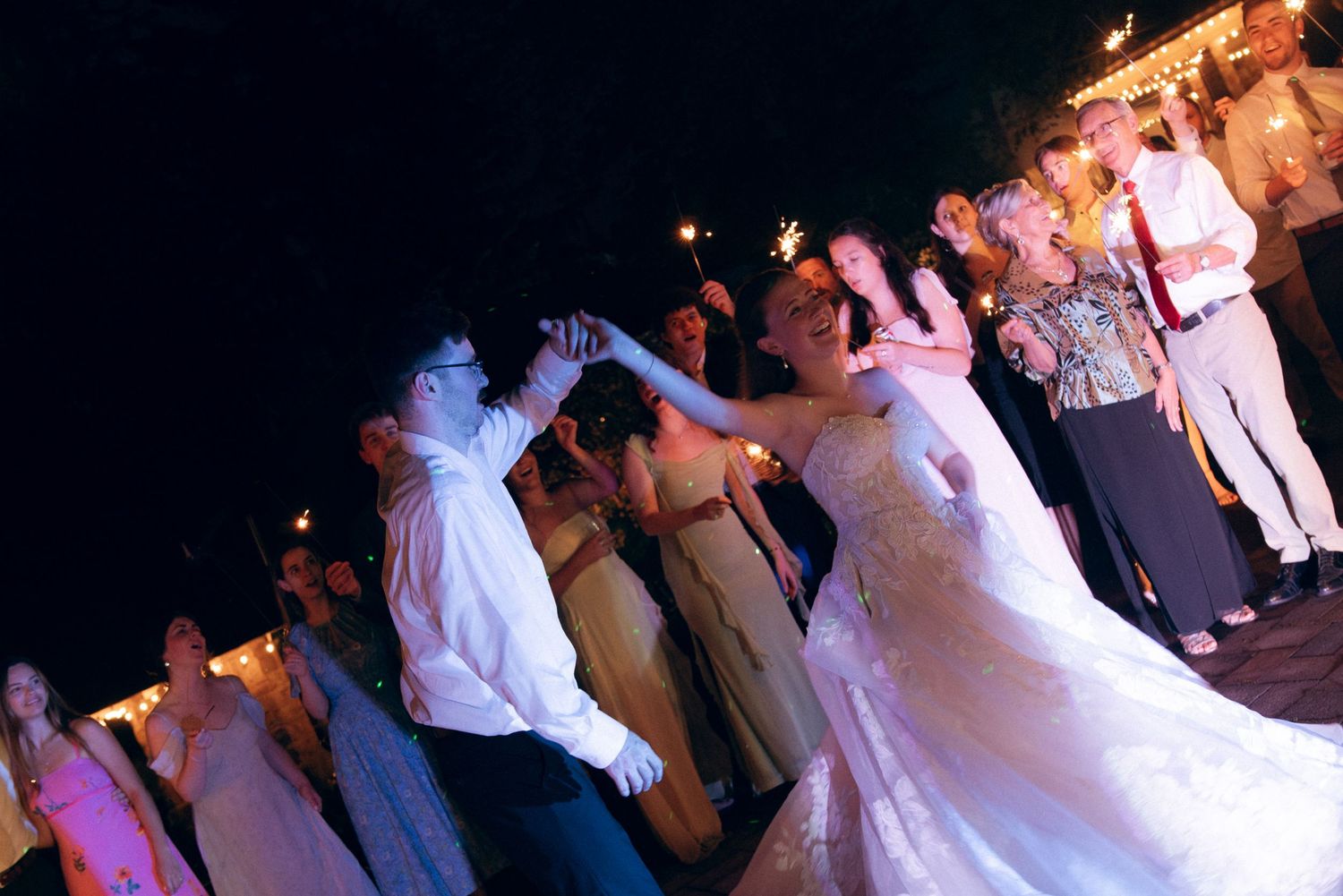 Couples dancing at an evening wedding reception with a crowd of people raising their arms in celebration against dark night sky.