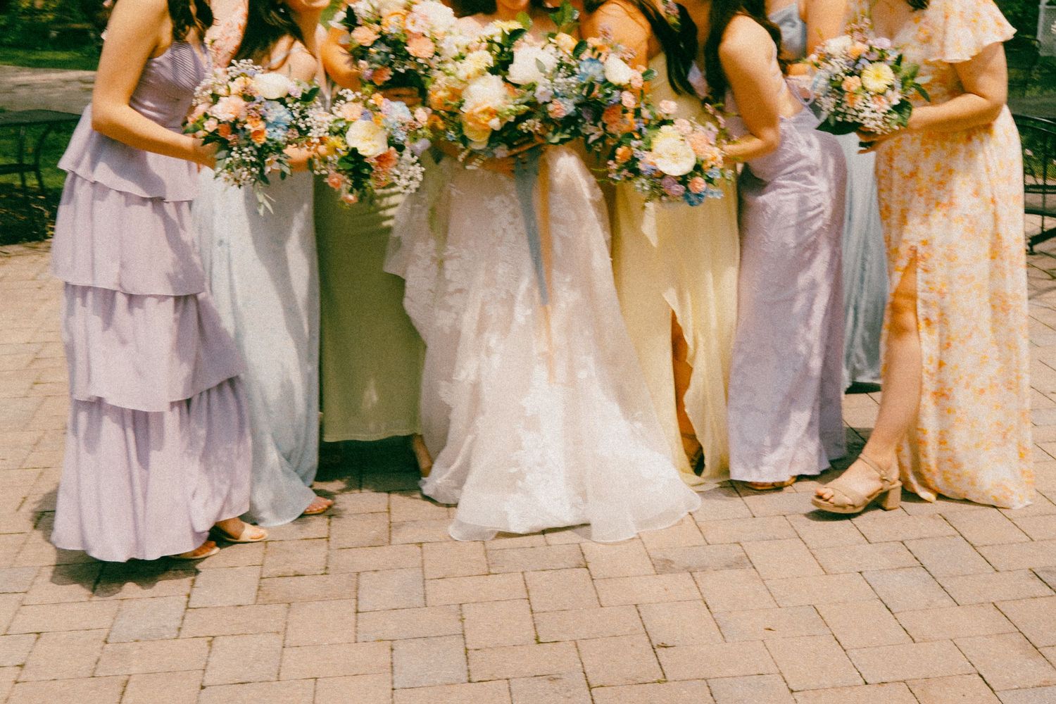 Bridesmaids in pastel dresses holding white and peach floral bouquets stand together on stone pavers outdoors.