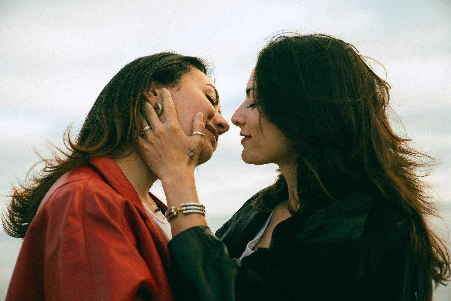 Two people share a tender moment together with their hair flowing in the breeze, one wearing red and one in black.