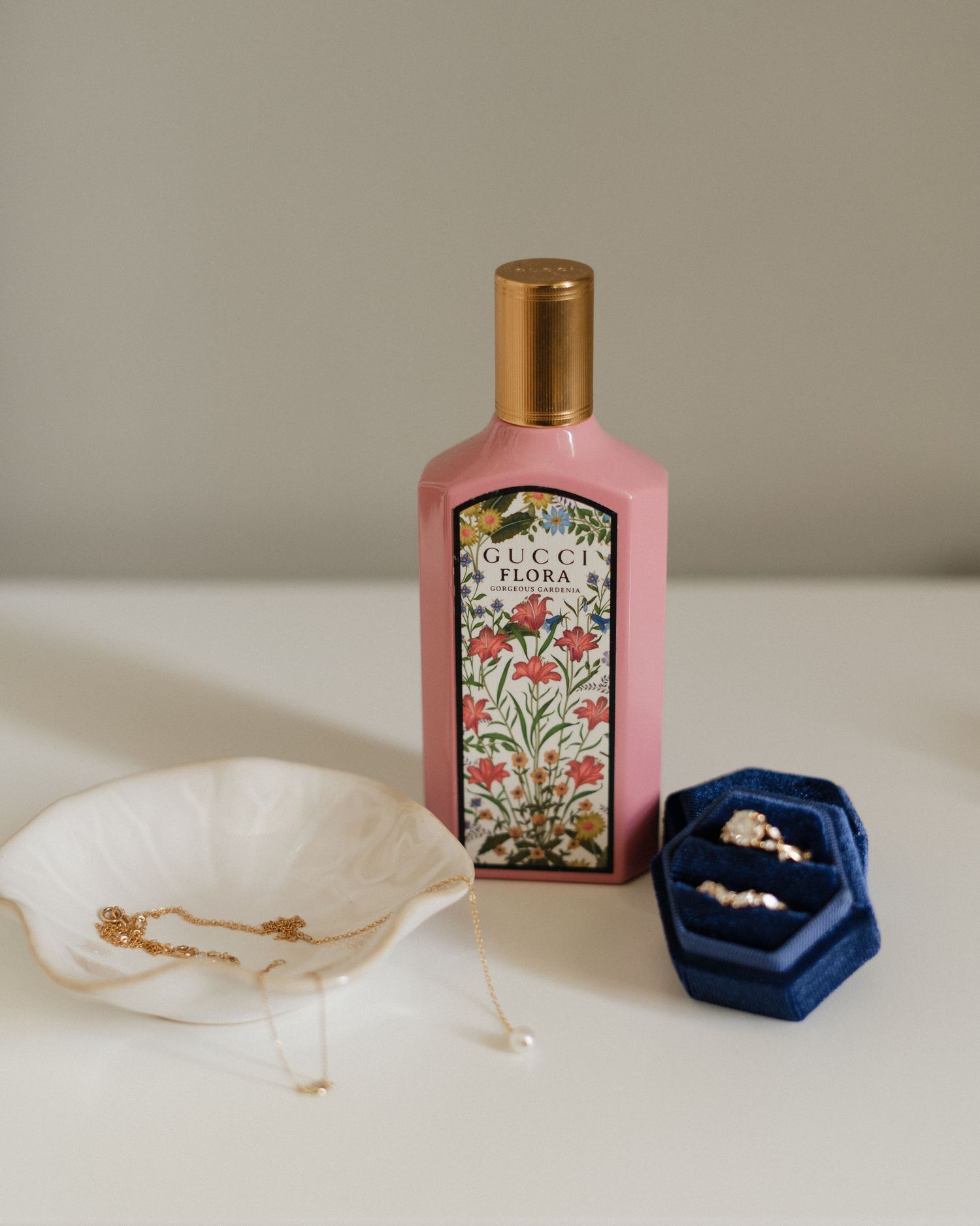 Pink perfume bottle with decorative floral design sits alongside a navy velvet ring box and crystal dish on white surface.