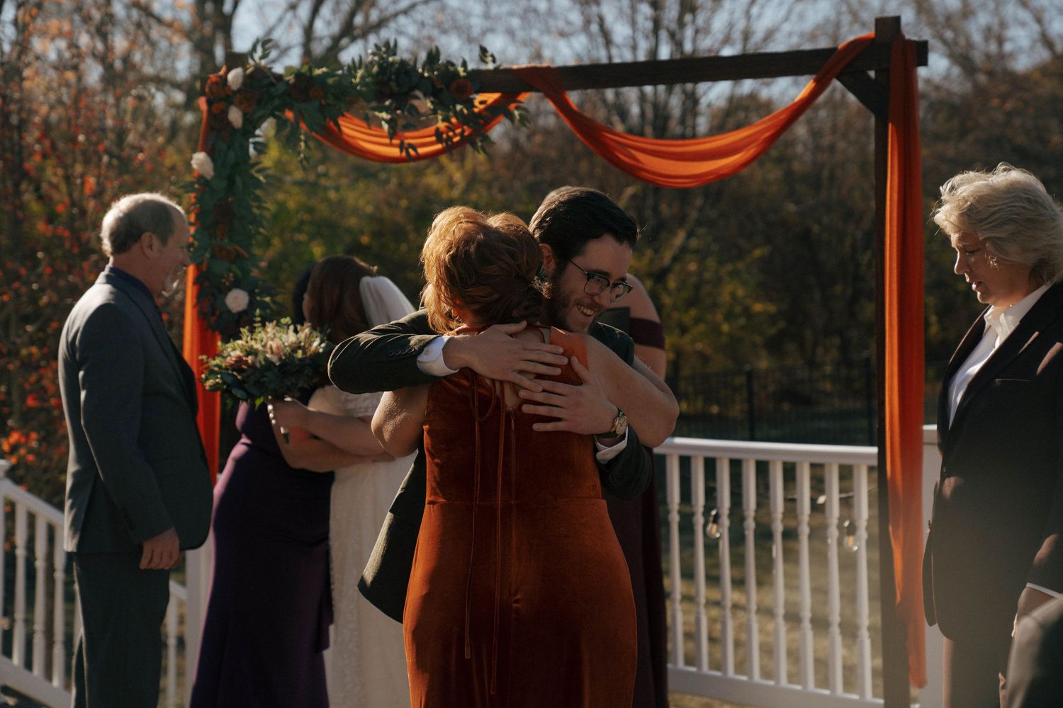 A tender moment during an autumn outdoor wedding ceremony beneath an orange-draped wooden arch.