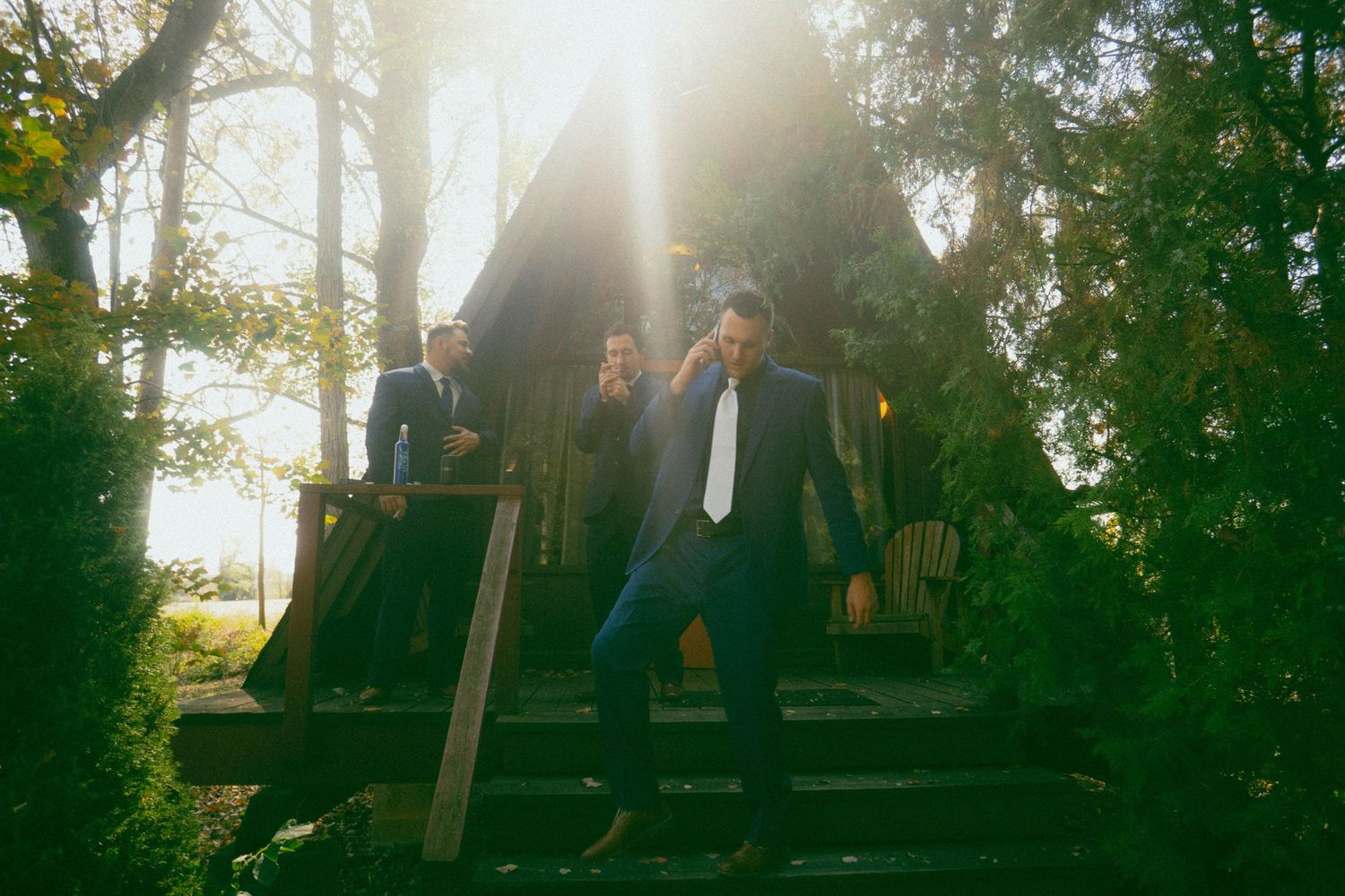 Two figures stand on wooden stairs backlit by golden sunlight through trees.