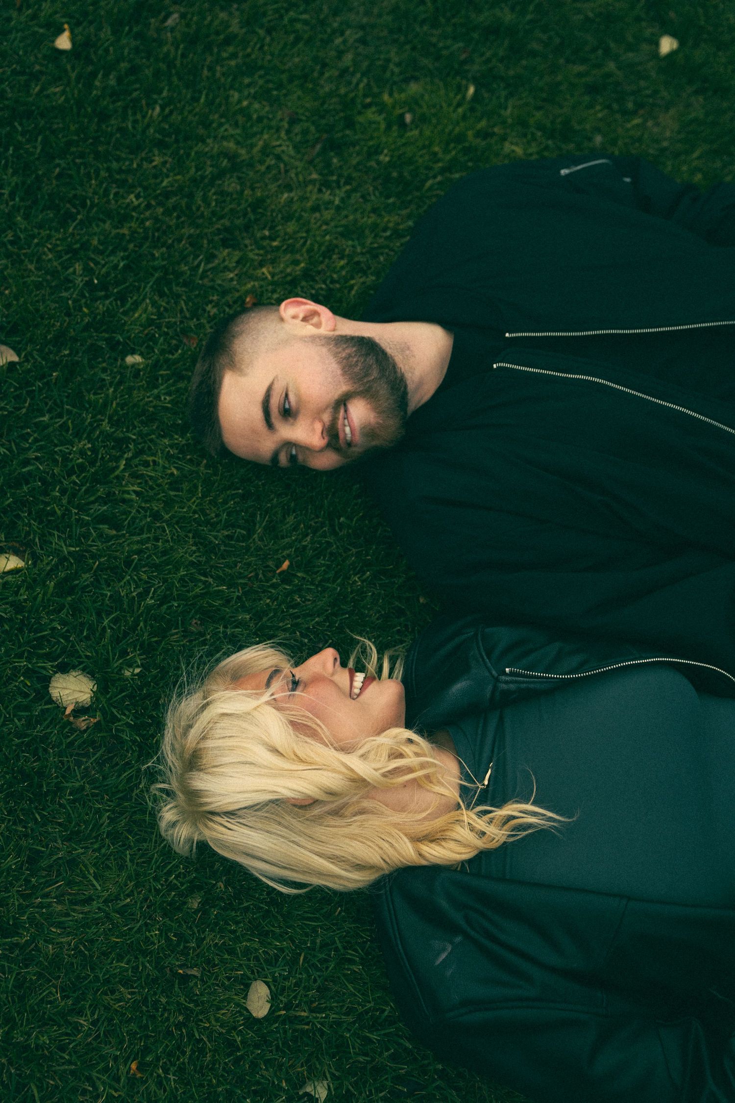 Person lying on grass wearing dark clothing with blonde hair spread out against dark green background. Couple in Bar Harbor Maine