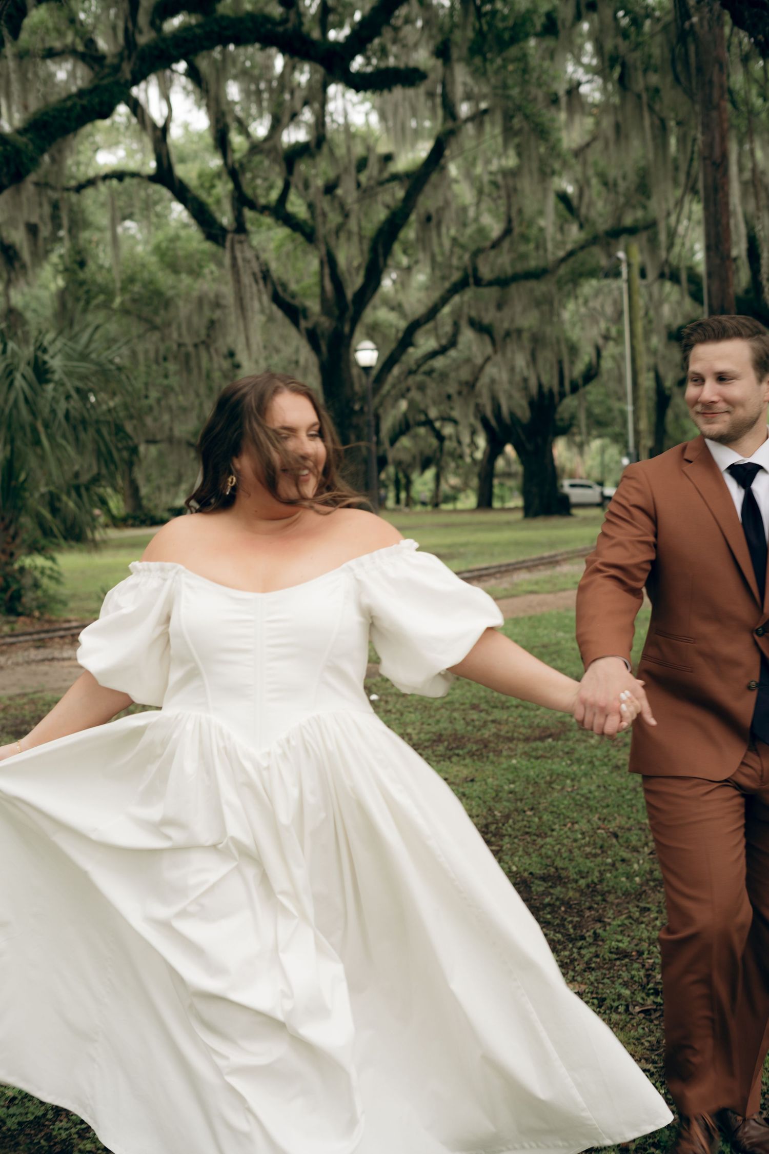 A couple in wedding attire spins playfully in a park setting with moss-draped oak trees in the background. New Orleans