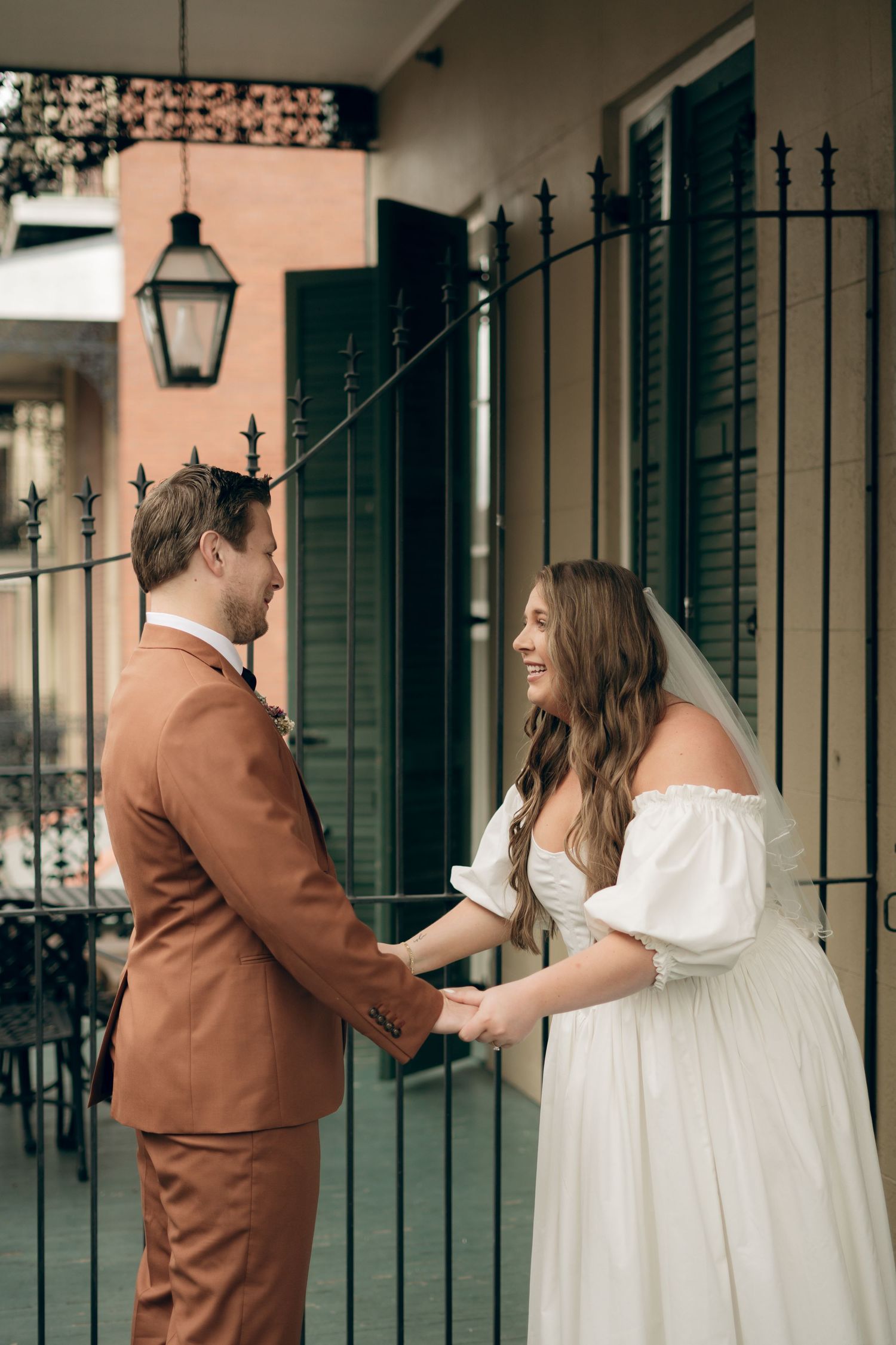 A couple shares a joyful moment in formal attire by a wrought iron gate with green shutters and a vintage lantern.