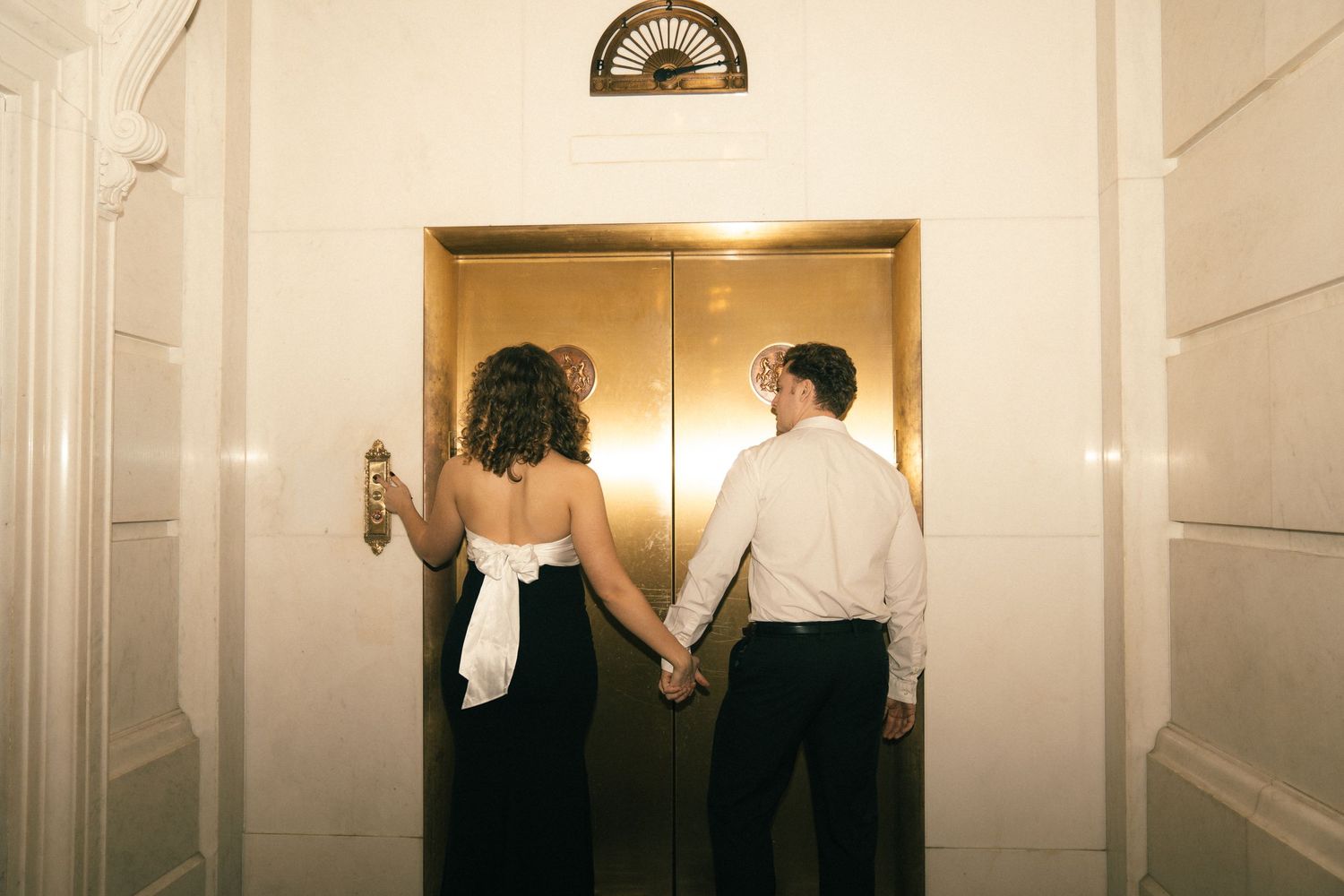 Two people in formal attire stand in an elegant hallway with white walls and a wooden door.