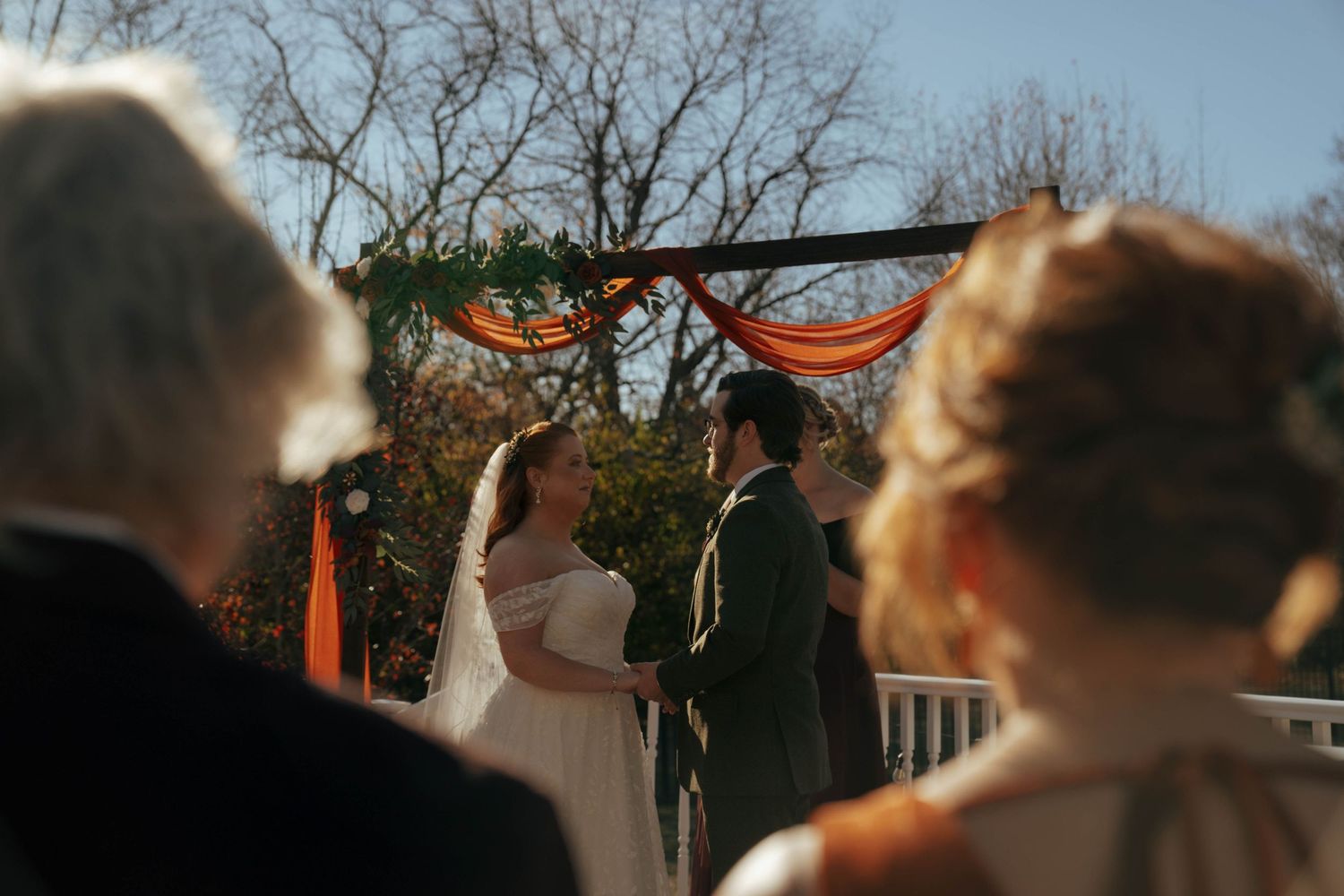 Wedding guests watch a ceremony under an orange-draped arch with sunlight filtering through trees.