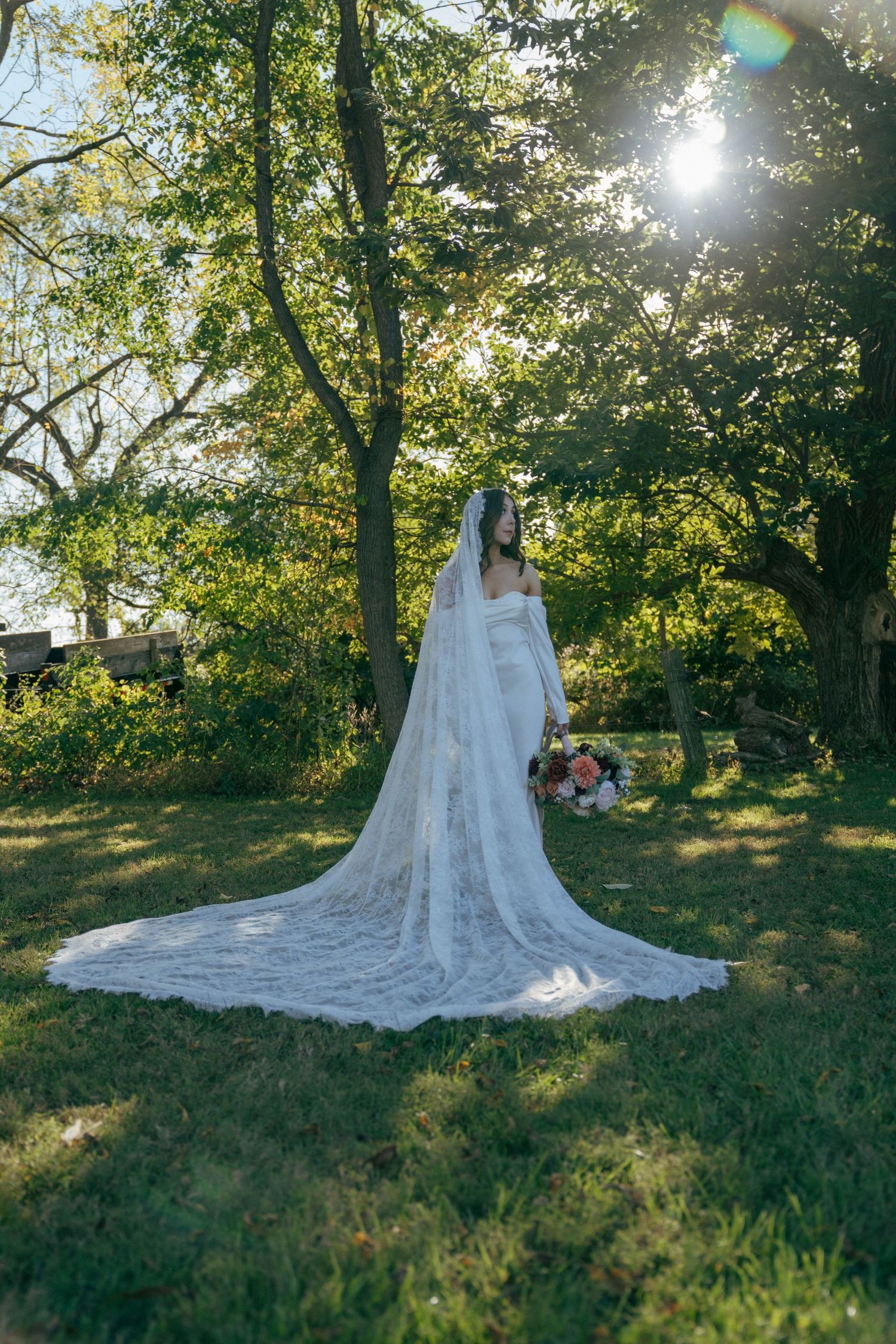 Bride in flowing cathedral veil stands in sunlit garden setting.