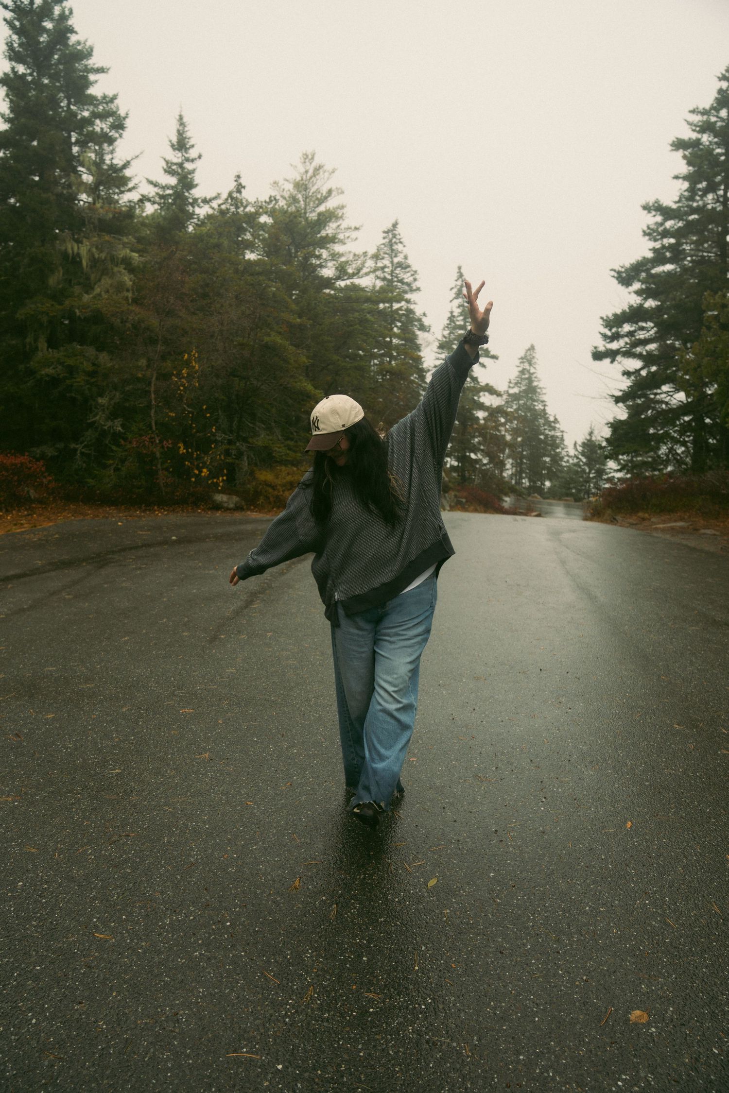A person in dark clothing walks on a wet road surrounded by evergreen trees on an overcast day.  Acadia National Park