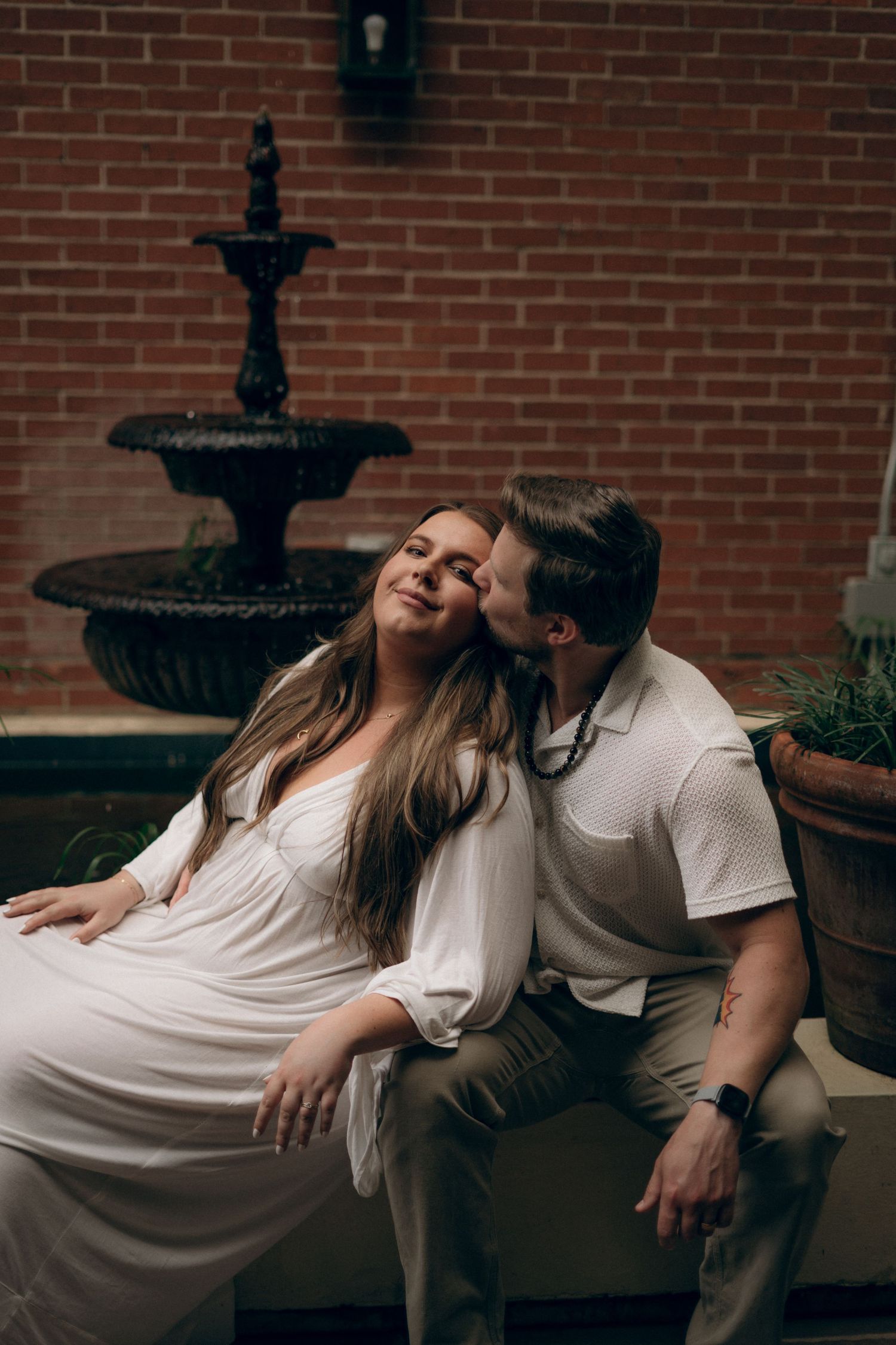 Romantic maternity couple relaxing on bench against brick wall with decorative fountain.